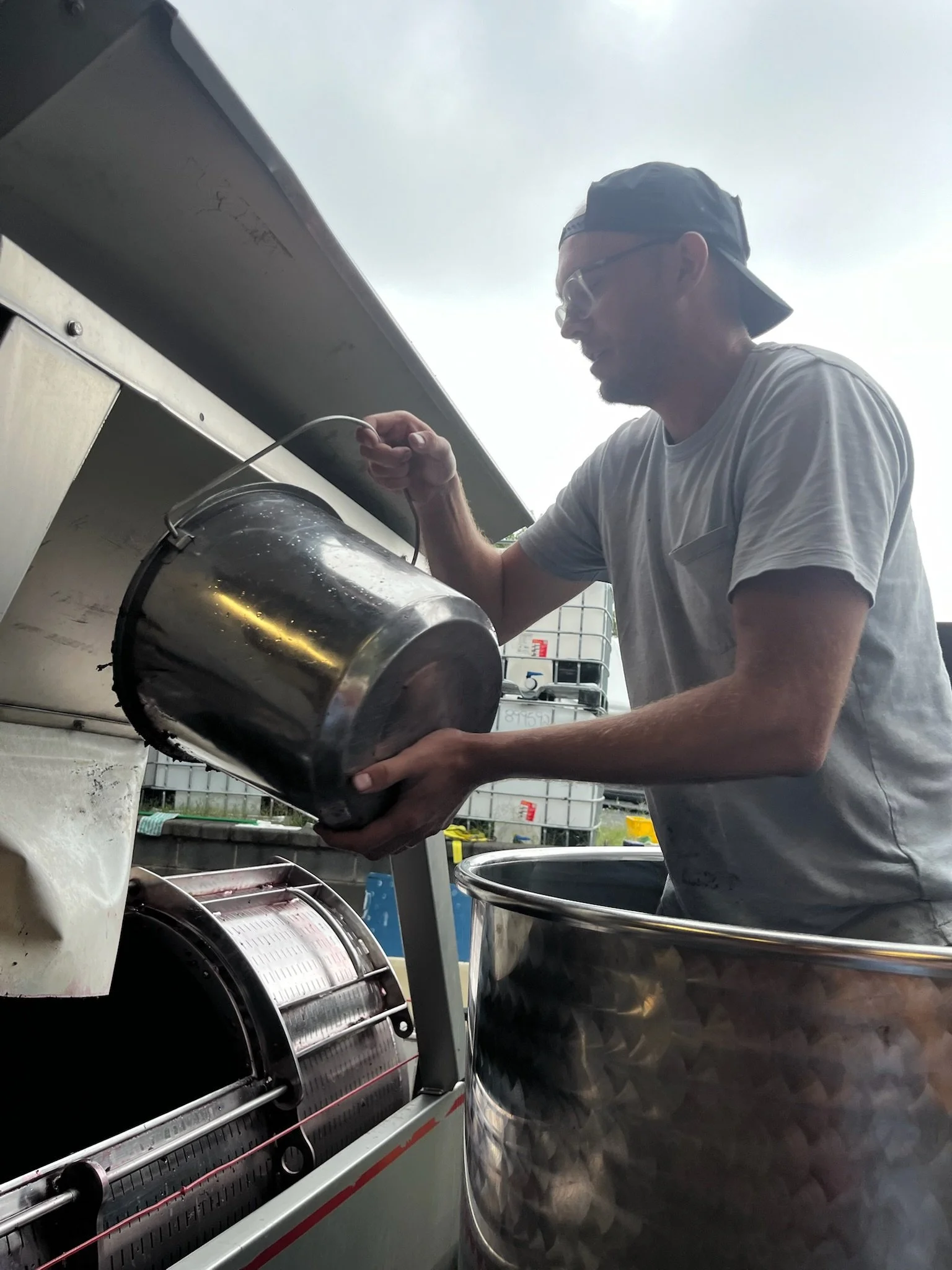 A man wearing a cap backwards, glasses, and a gray t-shirt is loading a large metal container into a commercial ice shaver machine outdoors. There are containers and equipment in the background.