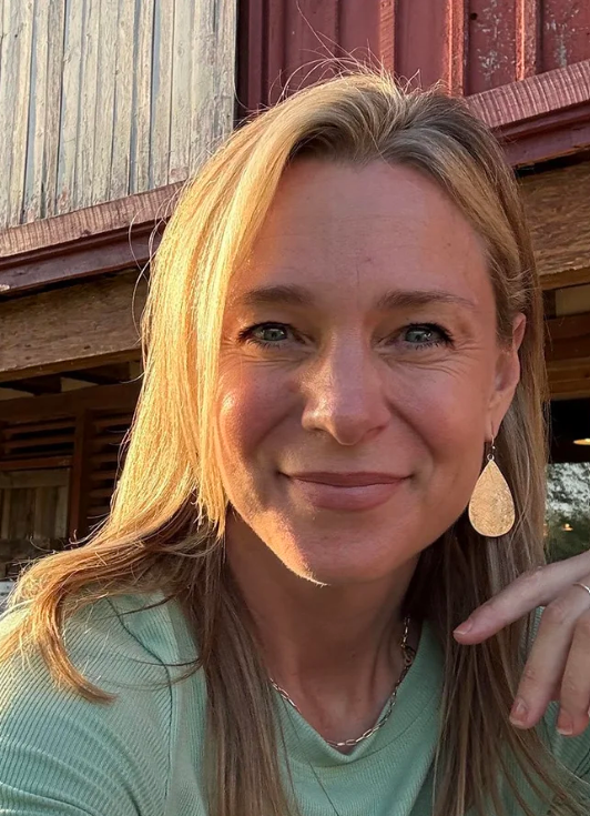 A woman with blonde hair and blue eyes smiling outdoors.