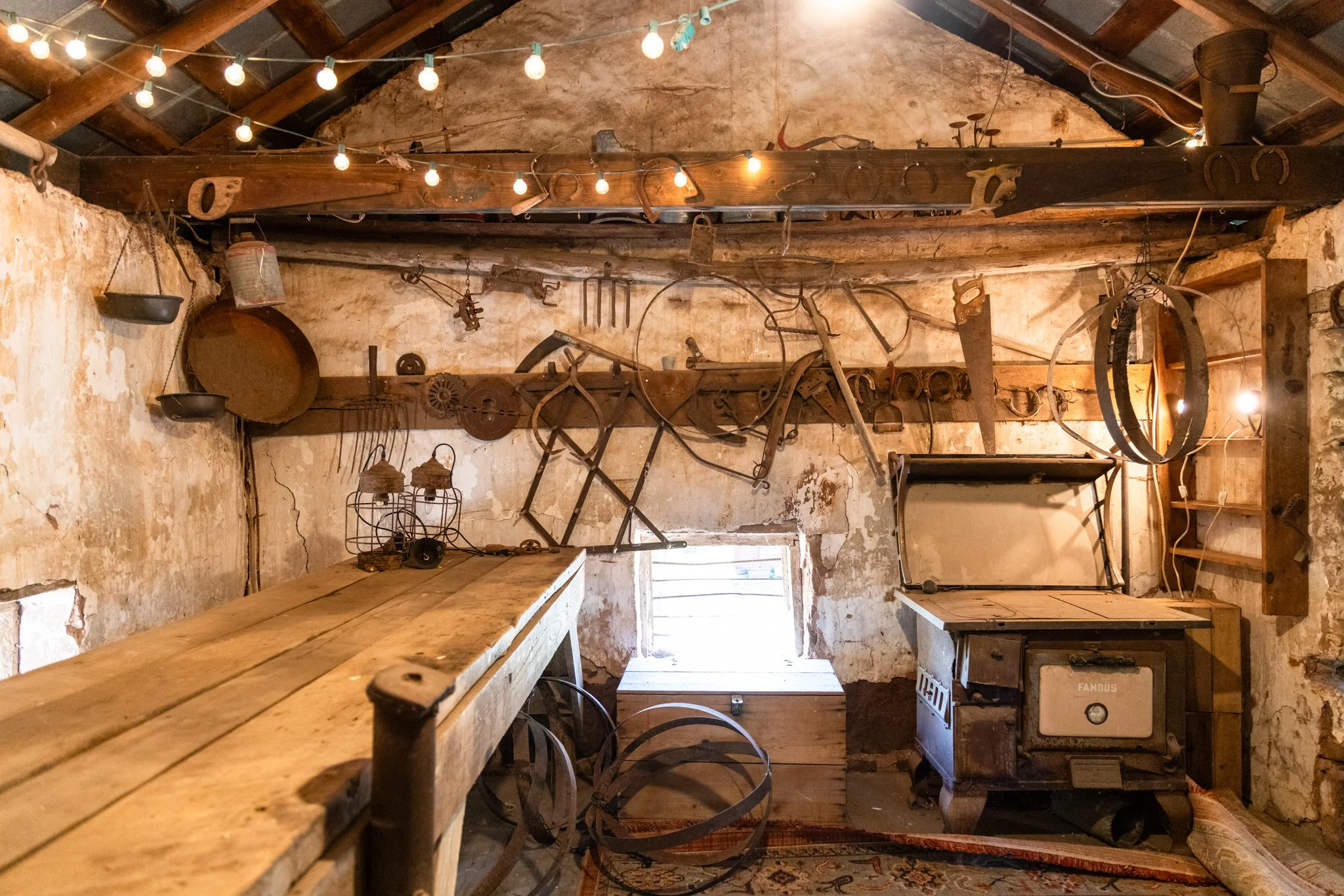 Interior of a rustic room with wooden beams, brick walls, and vintage tools hanging from the ceiling and walls, including horseshoes, a lantern, and cooking utensils. String lights are visible across the ceiling.