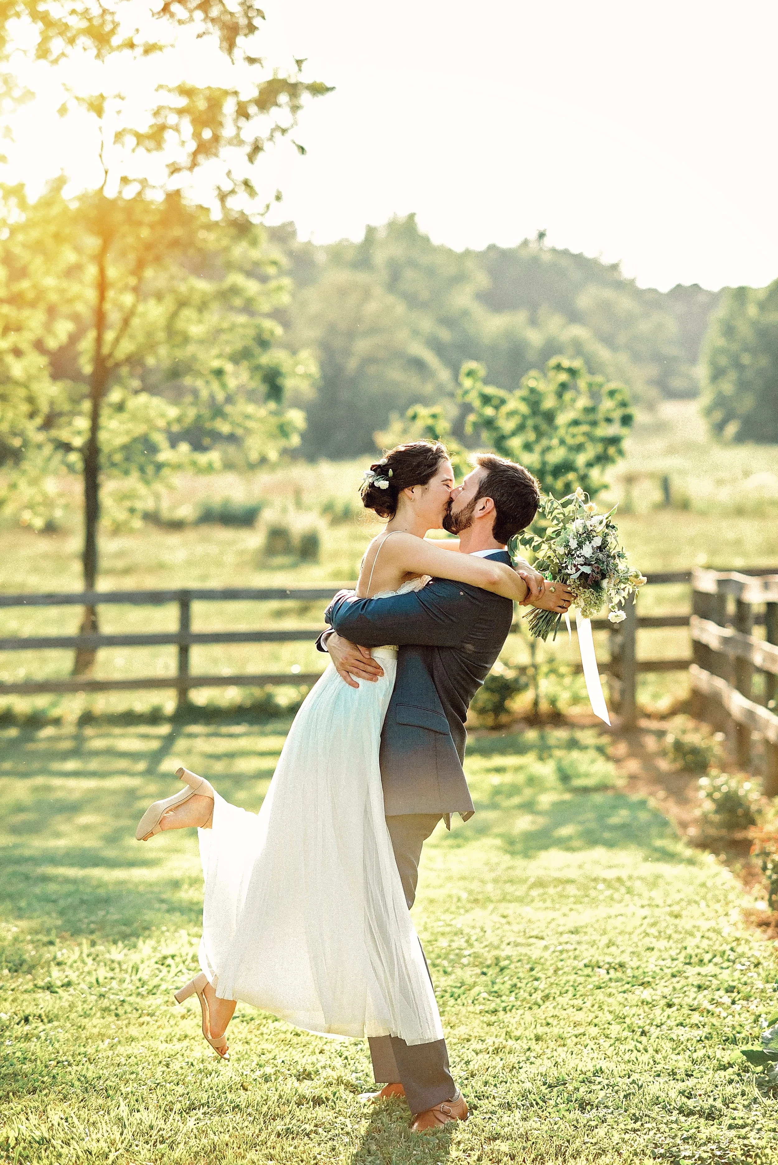 A newlywed couple is outdoors, with the groom lifting the bride in his arms after their wedding ceremony at Rocklands Farm Winery