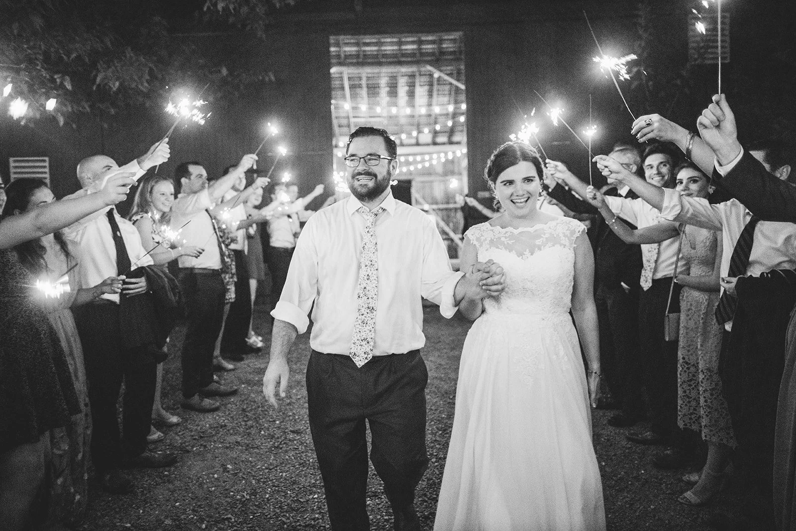 Black and white photo of a bride and groom walking hand in hand through a crowd of people holding sparklers during a nighttime celebration.