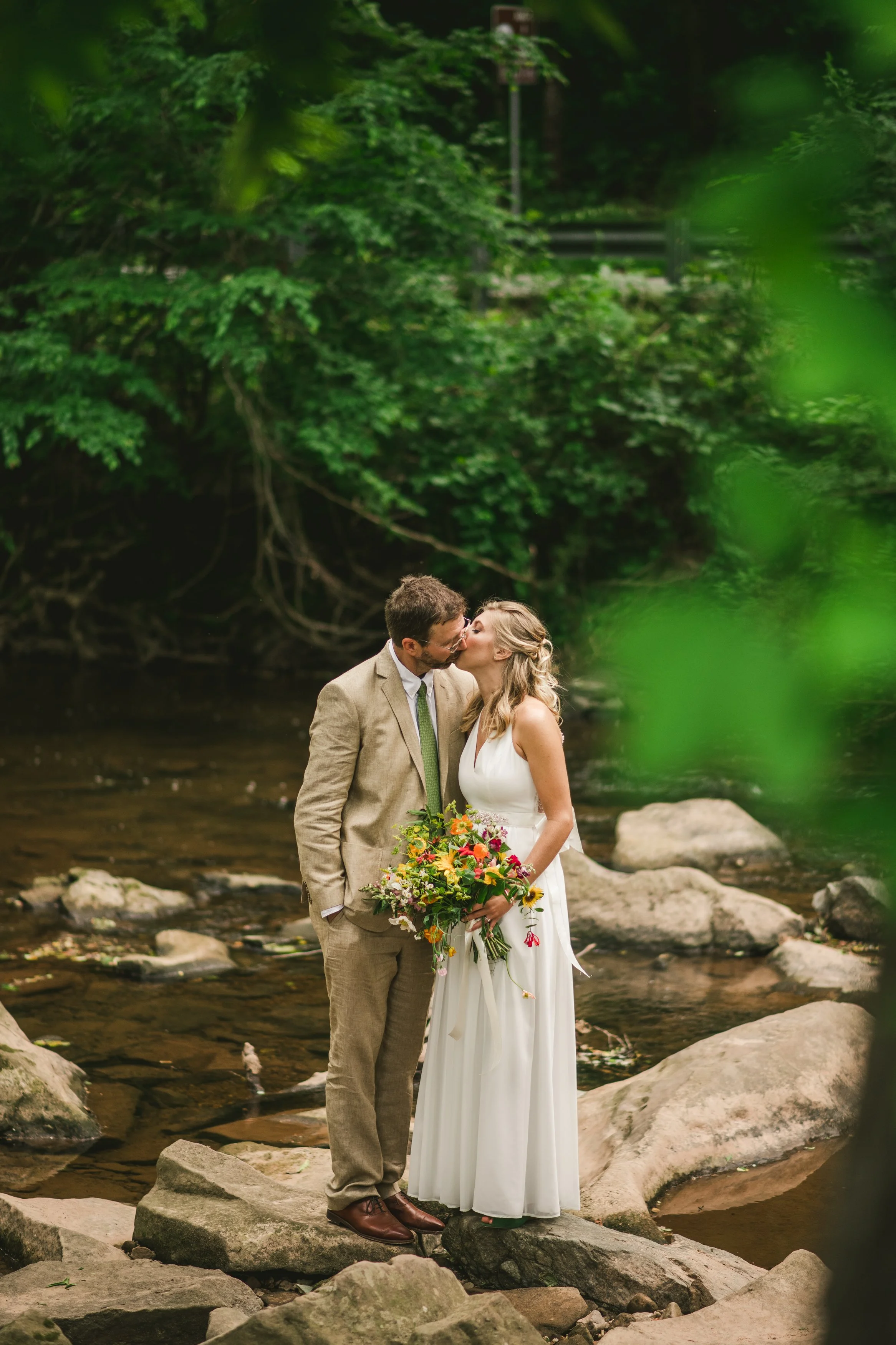 A bride and groom stand on rocks in a stream, sharing a kiss, with green trees in the background. The bride holds a bouquet of colorful flowers.