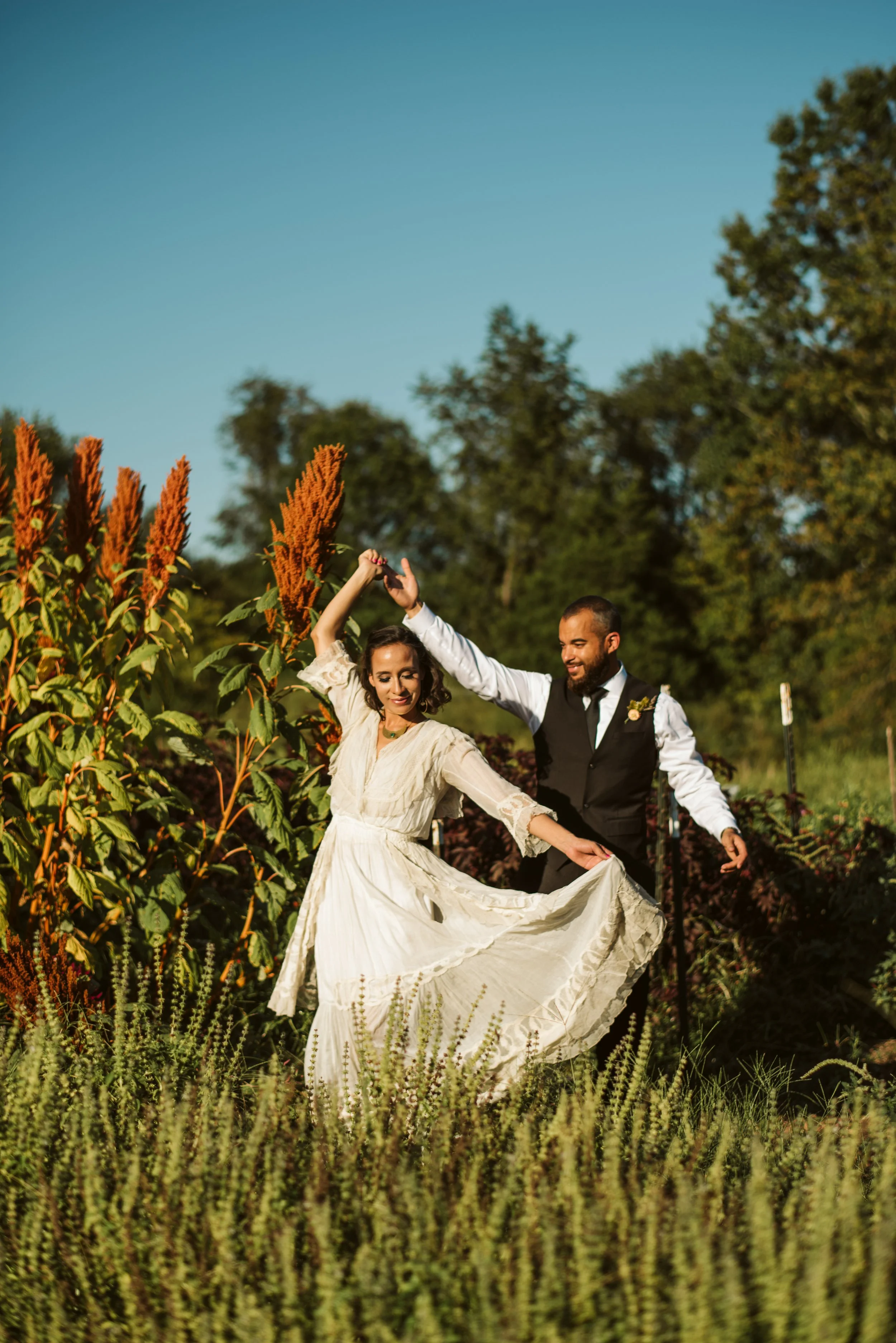 A couple dancing outdoors in a garden with tall plants and trees, under a clear blue sky during daylight.