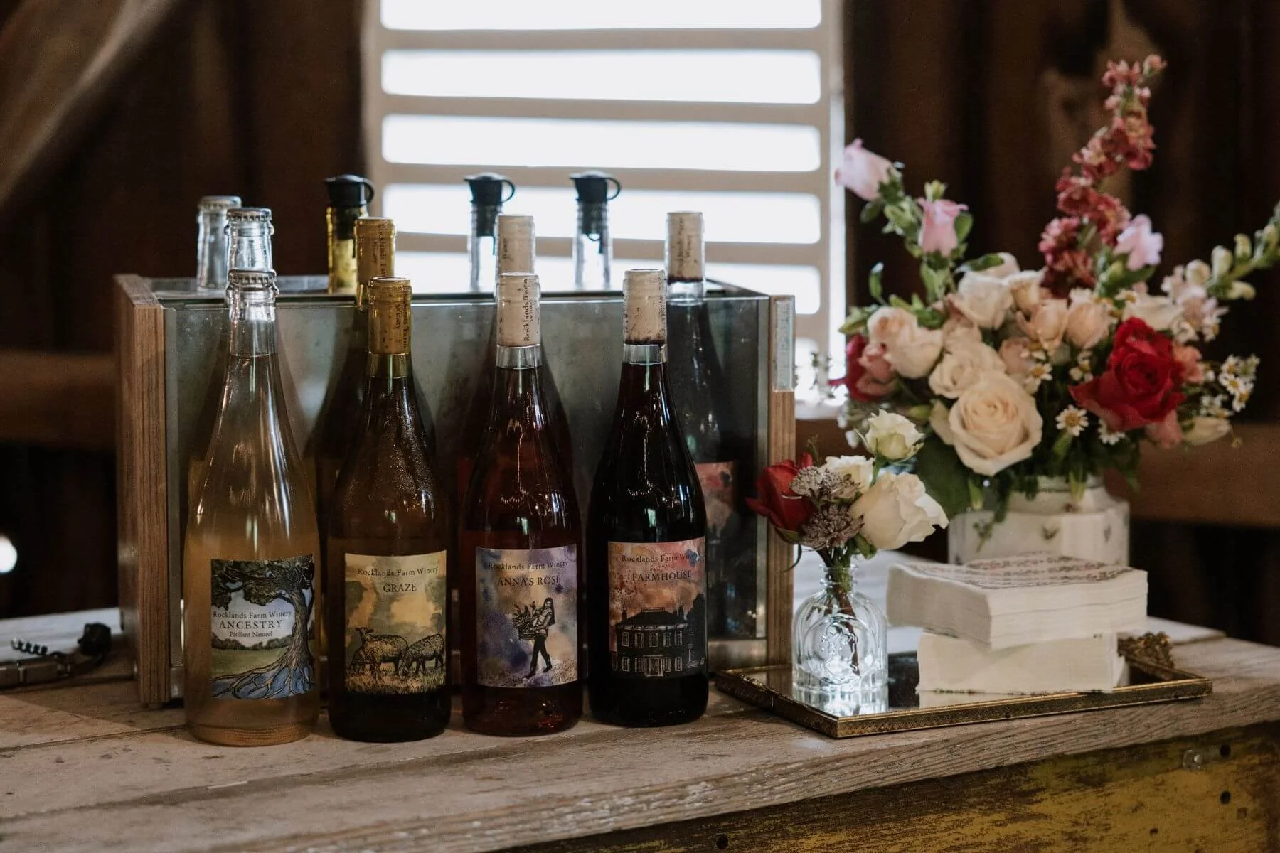 A display of four wine bottles with artistic labels, a glass vase with pink and white flowers, and a stack of napkins on a wooden surface inside a rustic setting with a wooden wall and window in the background.