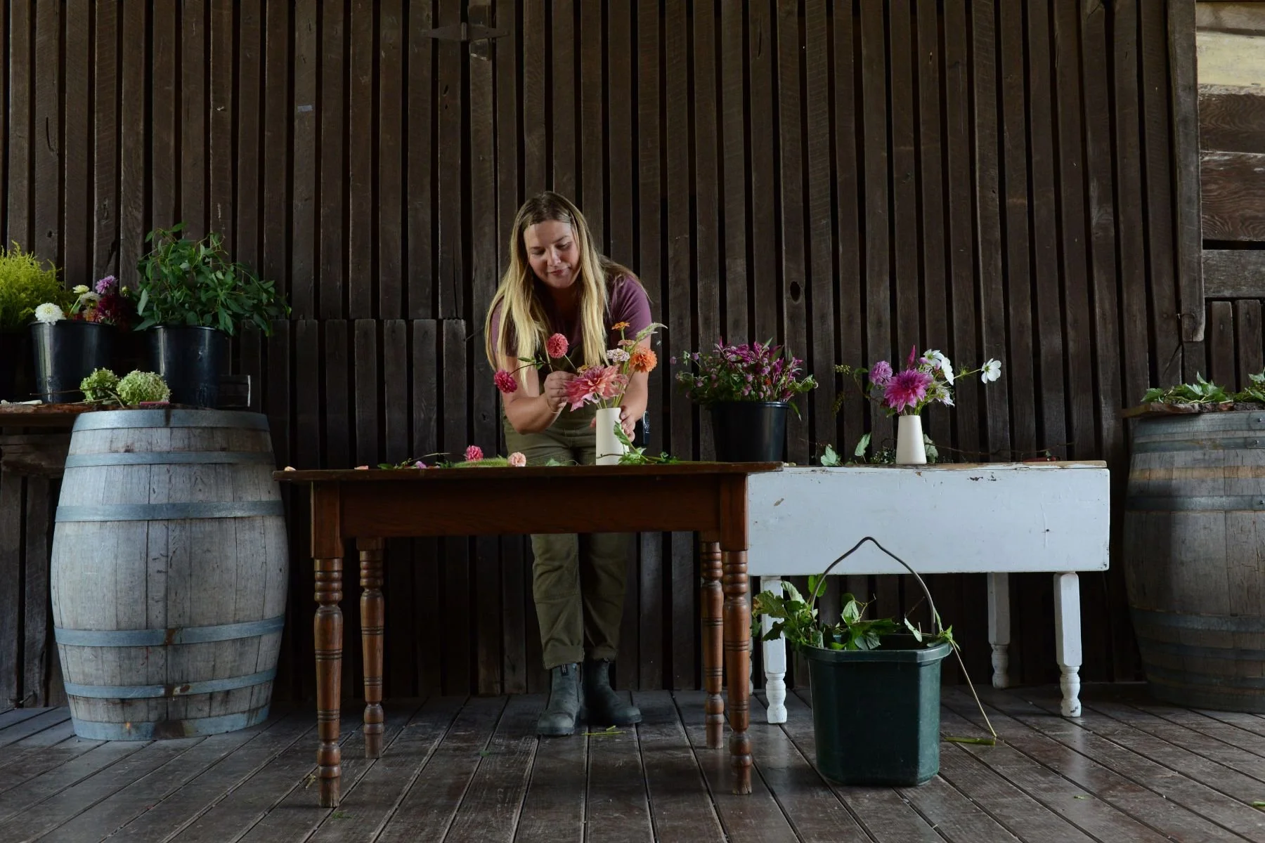 A woman arranging pink and purple flowers in a small white vase on a wooden table, with other flower arrangements in black pots and a white table in the background, set indoors with wooden walls and barrels.
