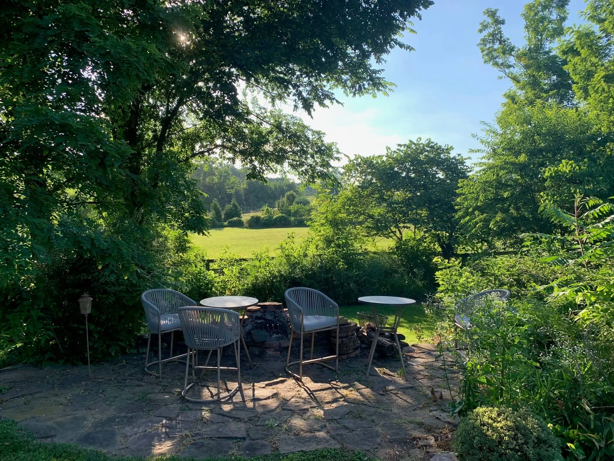 A backyard with a stone patio, white outdoor chairs around a fire pit, greenery, and trees under a blue sky with sunlight filtering through leaves.
