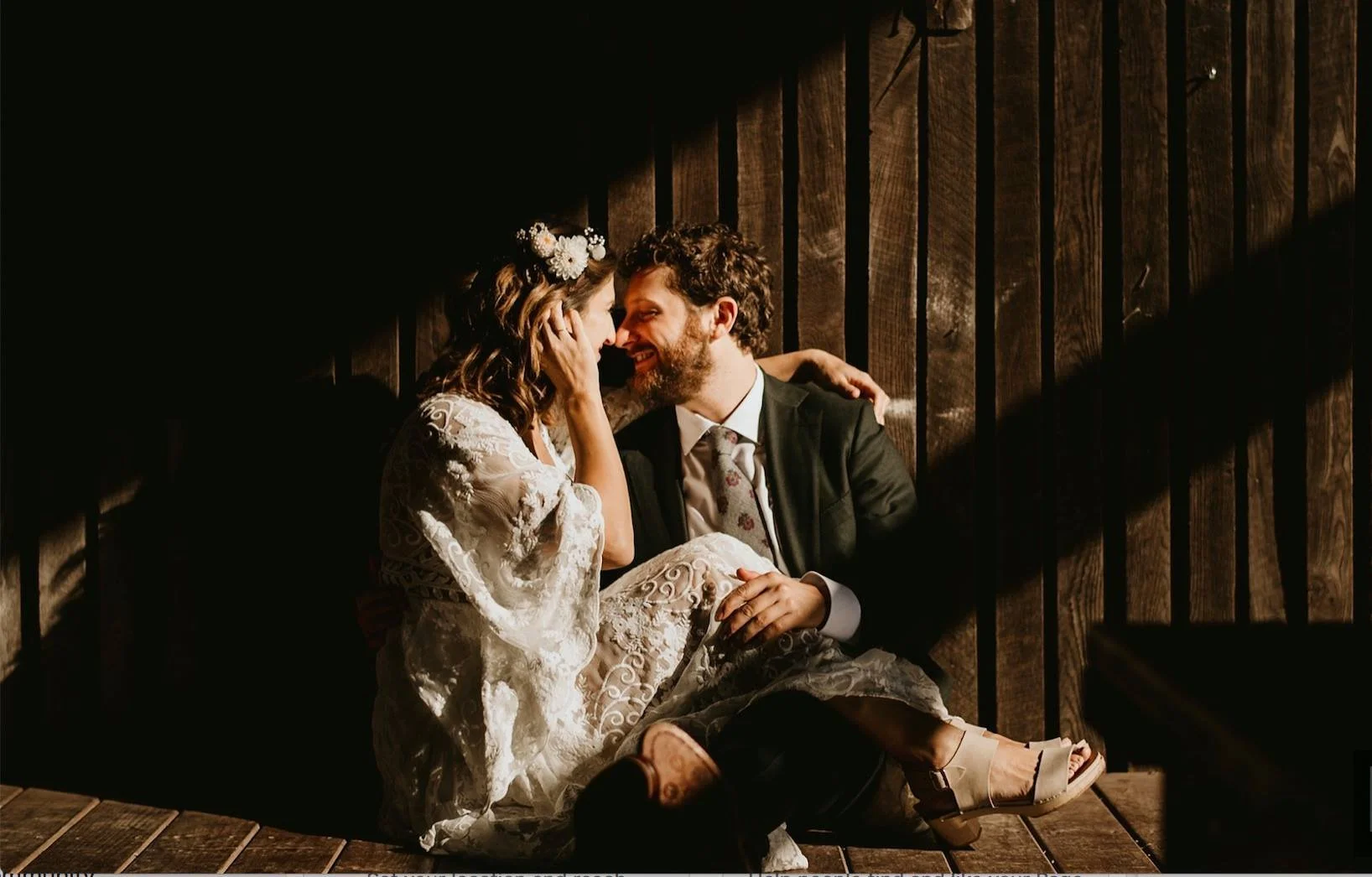 A couple sharing a joyful moment, sitting close together on a wooden floor with a dark wooden wall behind them, dressed in wedding attire.