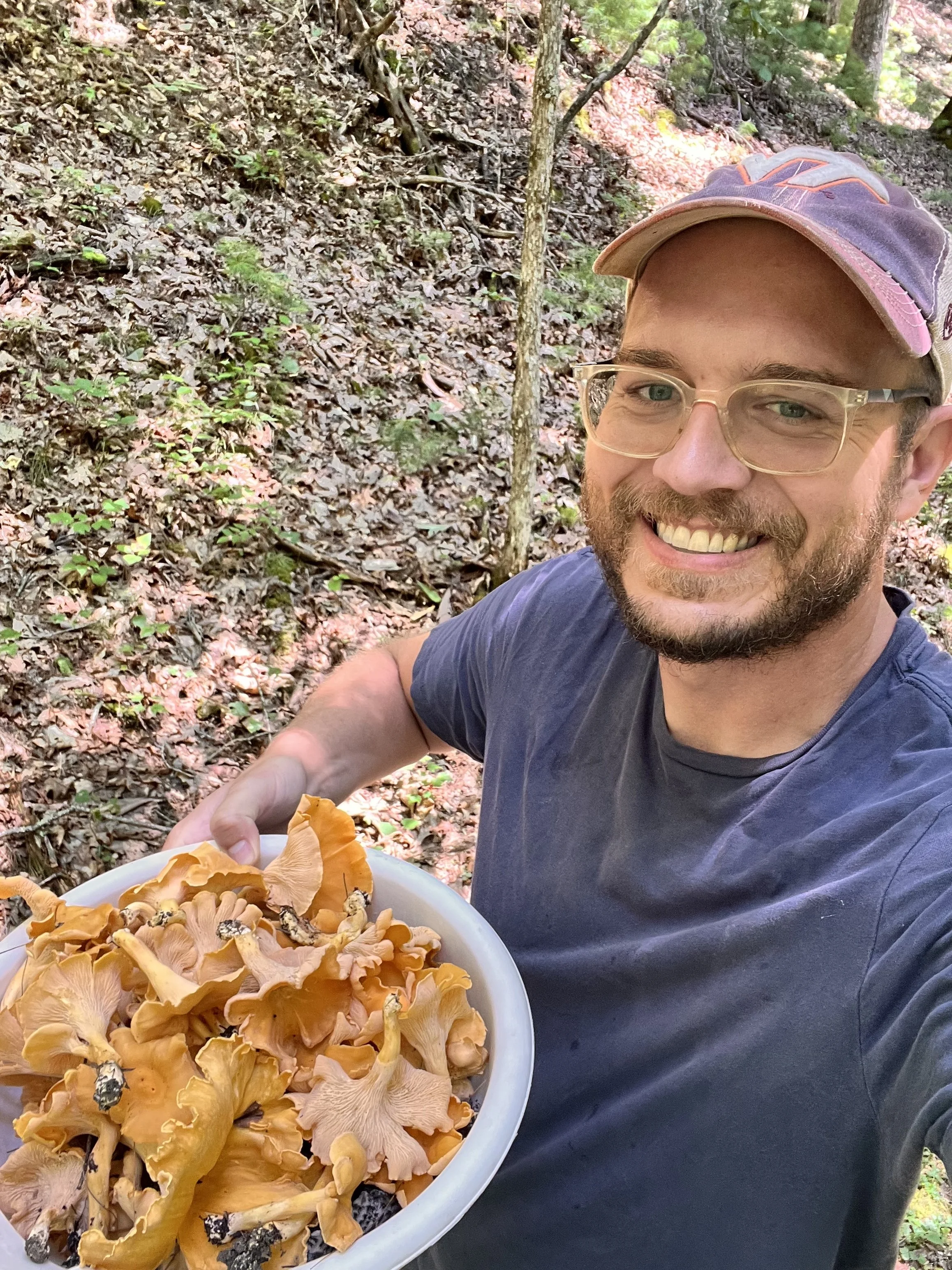A man outdoors in a wooded area, holding a bowl of freshly foraged wild mushrooms.