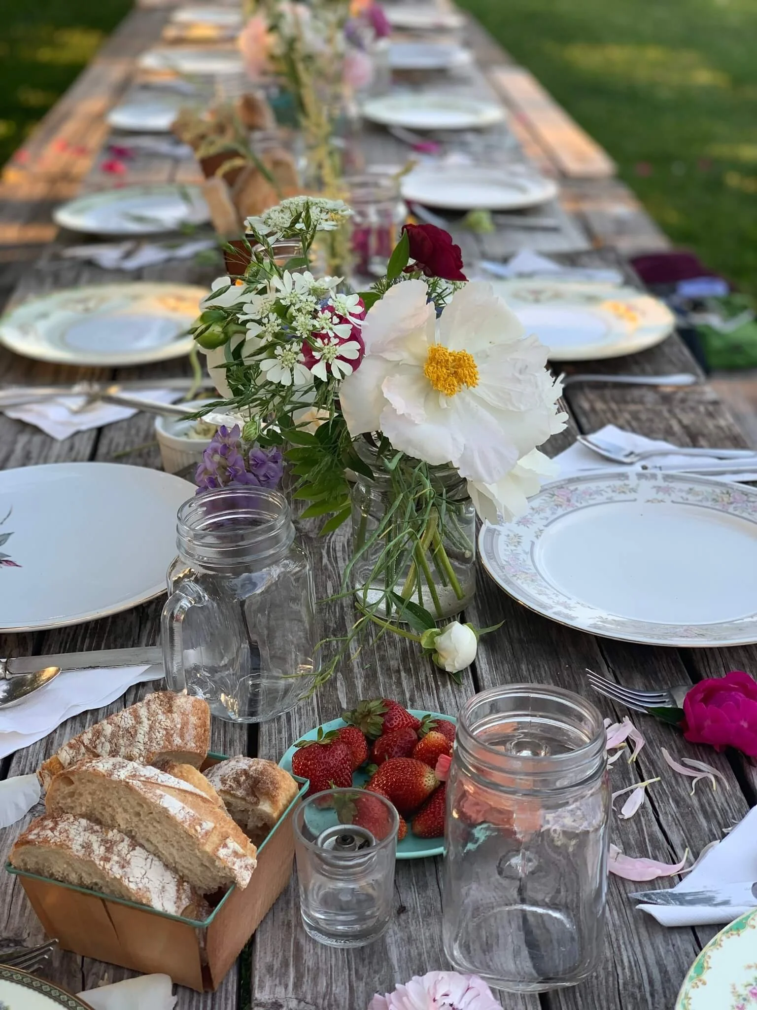 A rustic outdoor table set for a gathering at Rocklands Farm Winery, decorated with a floral centerpiece featuring white and red flowers, surrounded by plates, cutlery, glasses, fresh strawberries, bread, and a jar of water. 