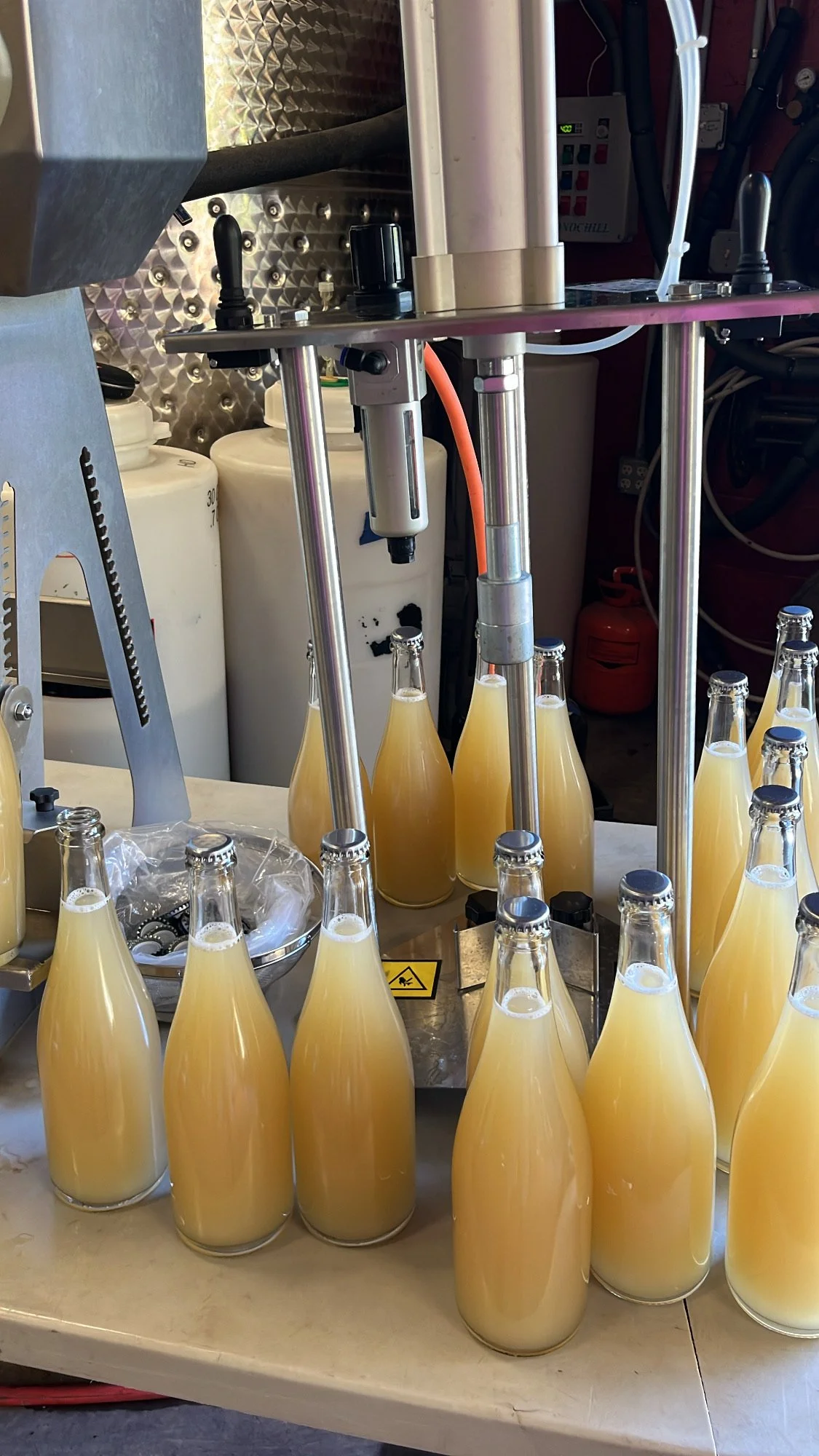 Bottles filled with a light yellow liquid, likely juice, being sealed or processed by a bottling machine in a factory or production setting.