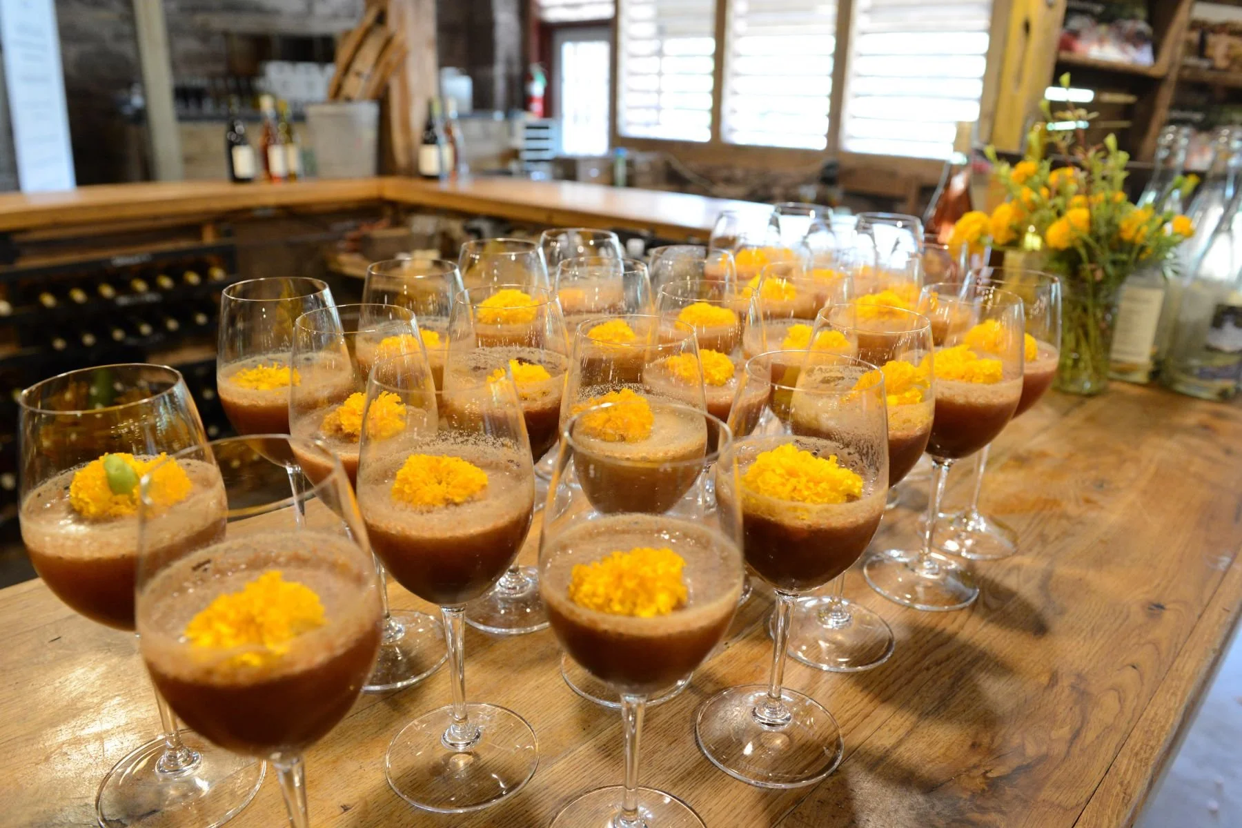 Several glasses of a brown-colored dessert topped with yellow flowers on a wooden table in a rustic setting, with flowers in a vase to the right.
