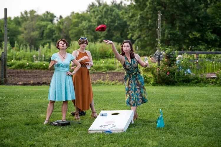 Three women playing outdoor bean bag toss game on a green lawn with trees and flowers in the background. One woman is throwing a bean bag, another is watching, and the third is holding a drink.