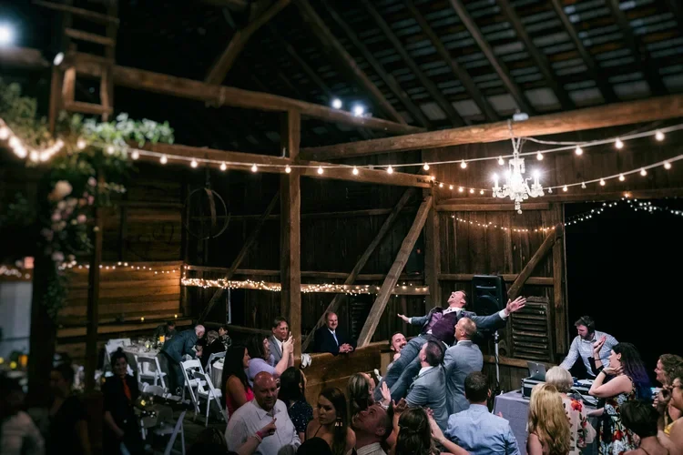 Guests dancing and celebrating at a wedding reception in a rustic barn decorated with string lights and a chandelier.