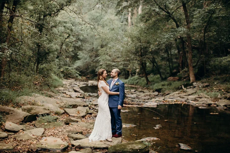 A bride and groom stand close together in a wooded area near a creek, sharing a romantic moment on their wedding day.