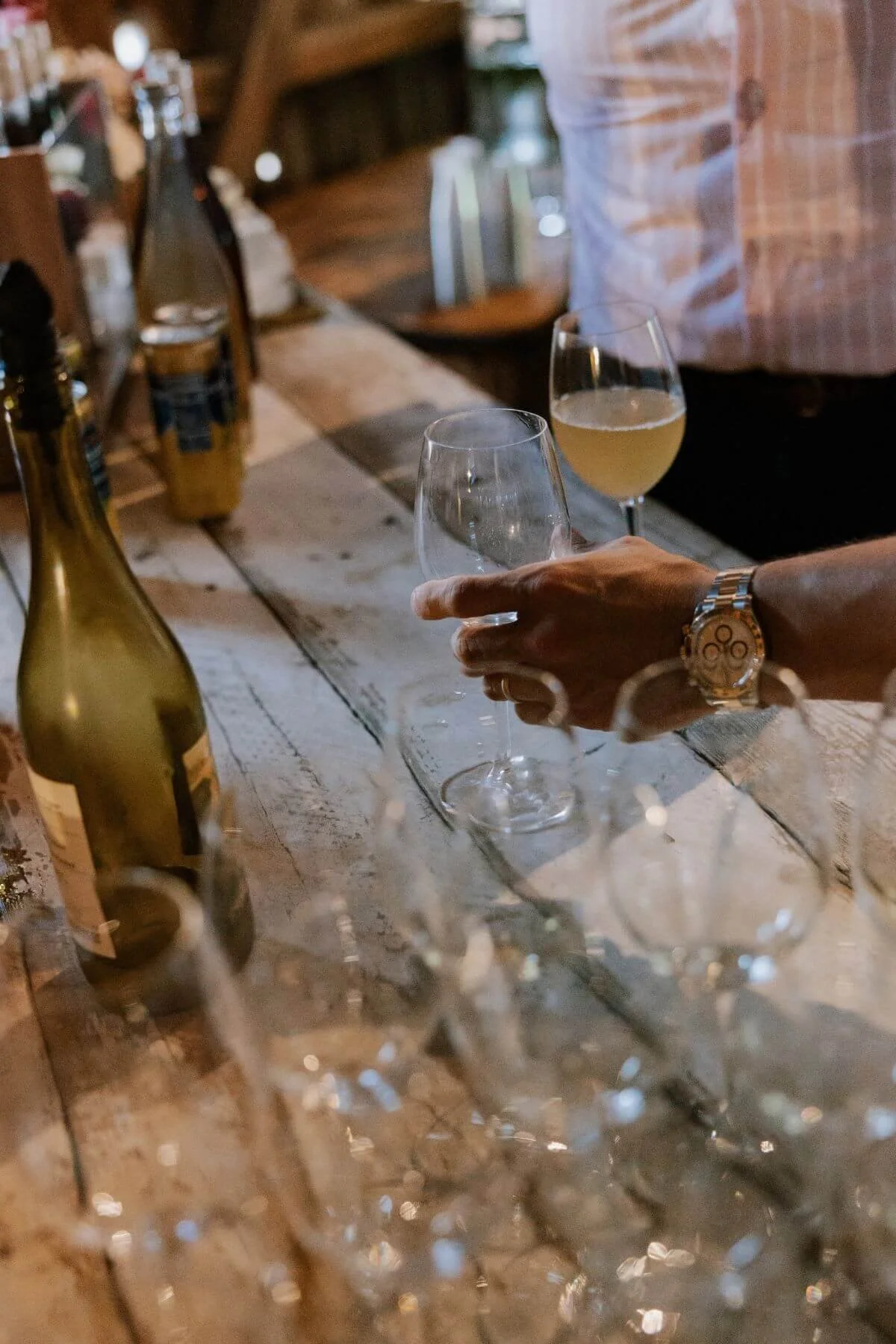 A person pours white wine into a wine glass at a rustic wooden bar, with bottles and barware in the background.