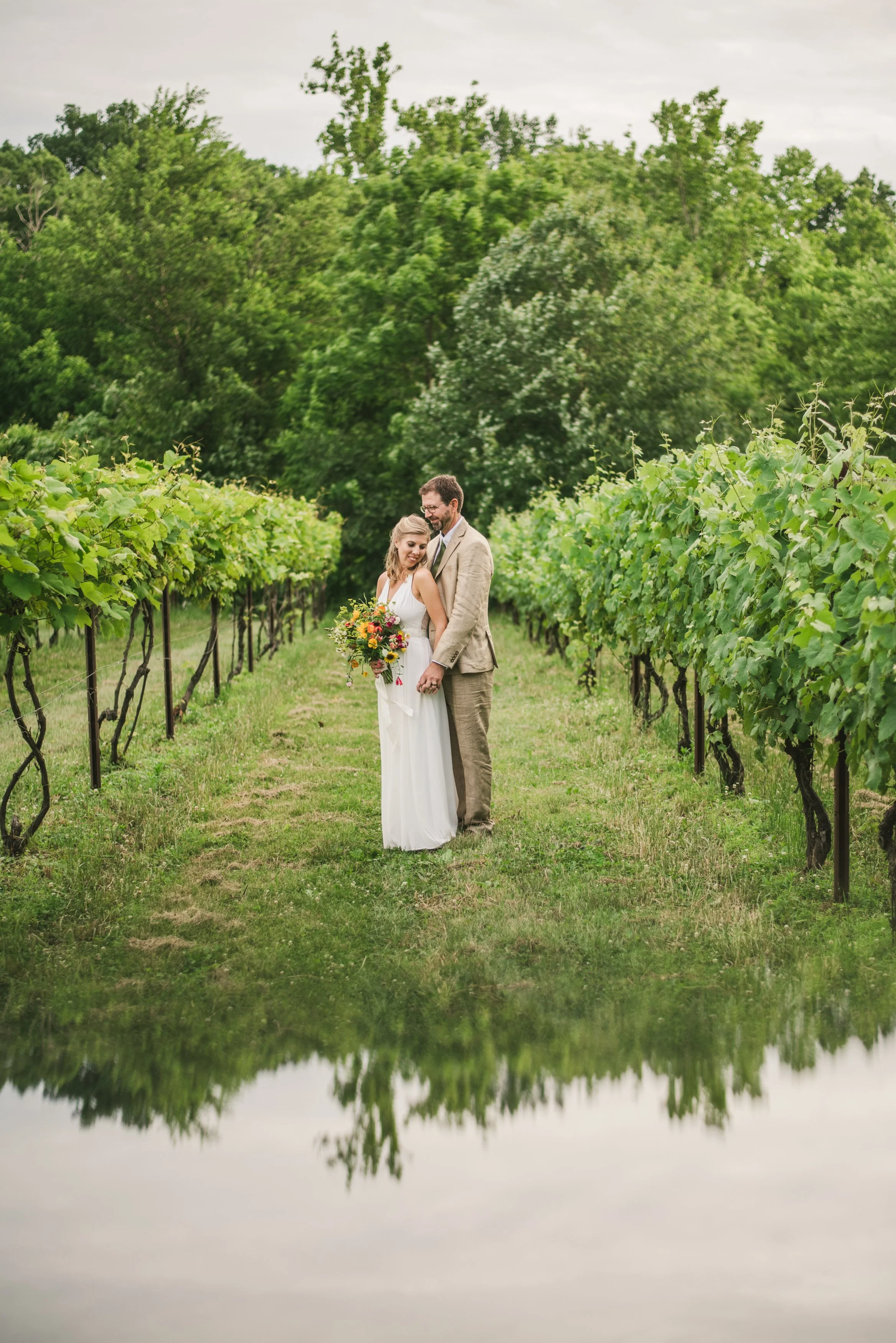 A couple stands in the vineyard at Rocklands Farm Winery, surrounded by lush green grapevines, with a large tree and cloudy sky in the background, and their reflection visible in a water mirror below.