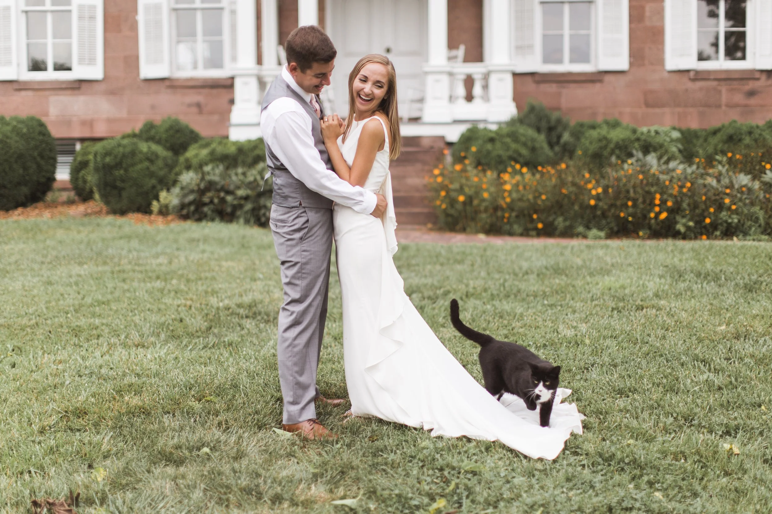 A bride and groom standing on grass in front of a brick house with white windows and a porch at Rocklands Farm Winery. They are smiling and holding each other, with a black and white cat on the ground near their feet.