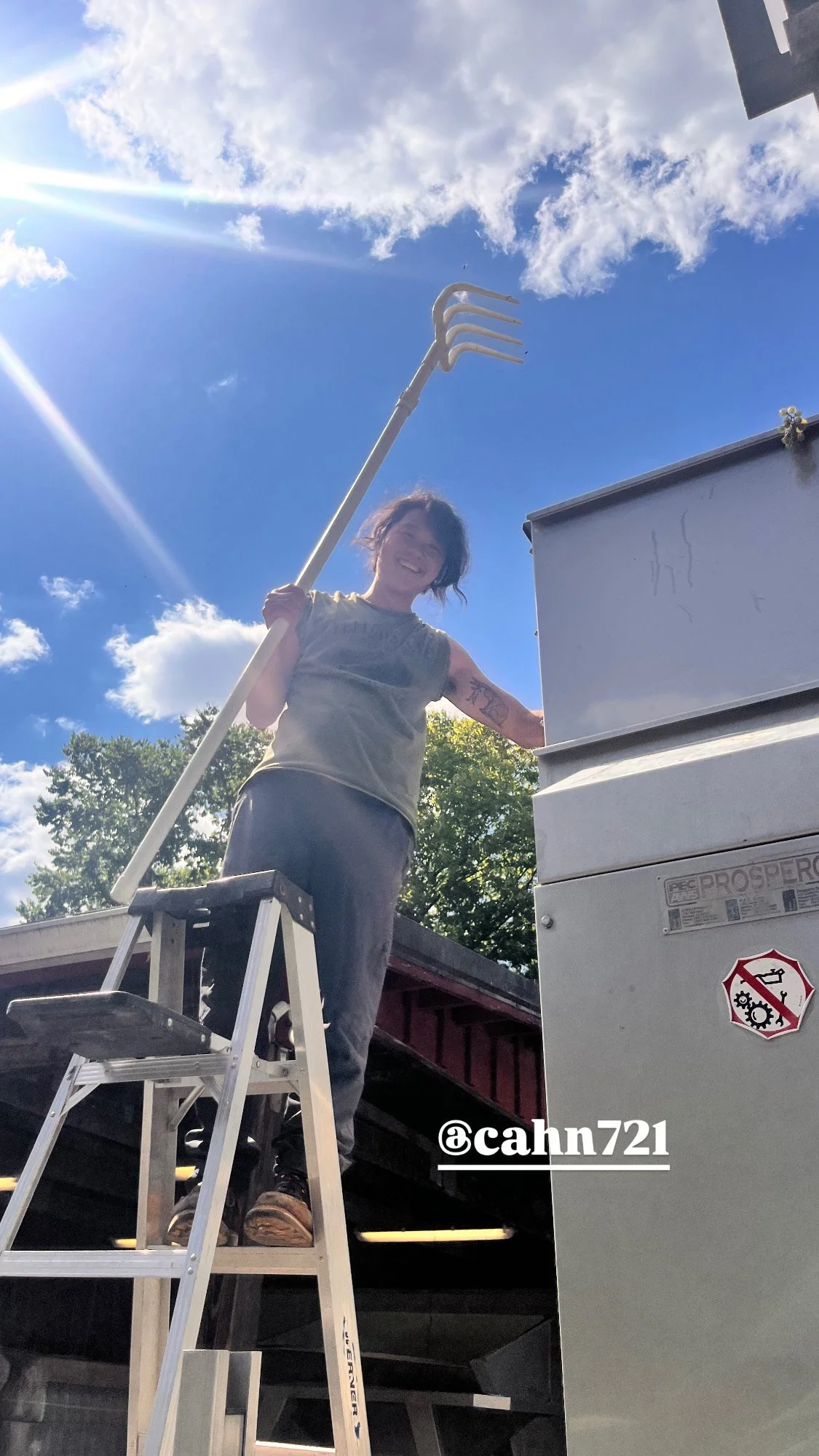 A smiling woman standing on a step ladder next to a commercial HVAC unit outdoors, holding a garden rake, under a partly cloudy sky.
