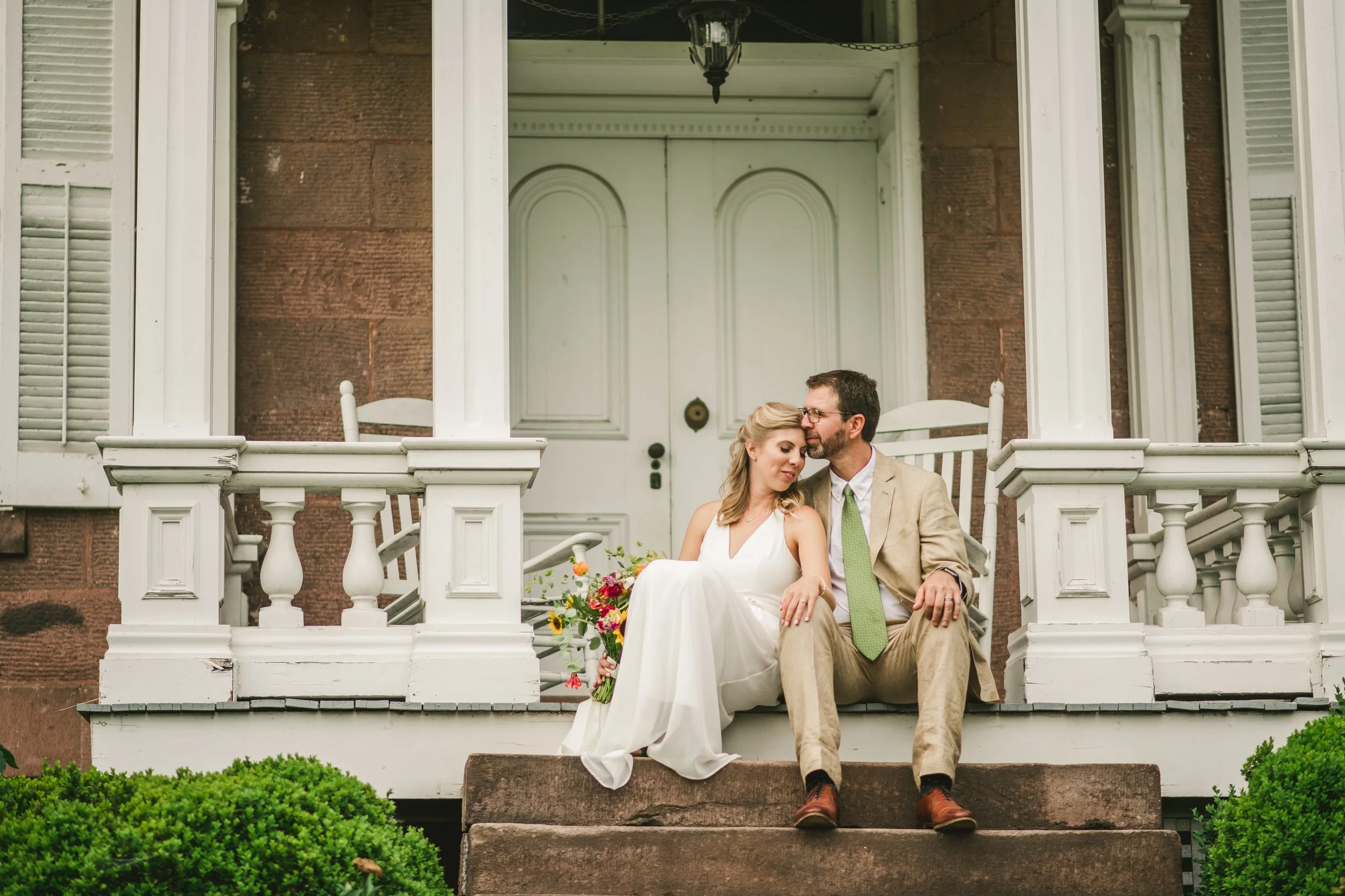 A couple sitting on the stairs of a front porch of a house, with the woman in a white wedding dress holding a bouquet, and the man in a tan suit with a green tie, leaning his head against hers as they sit close together.