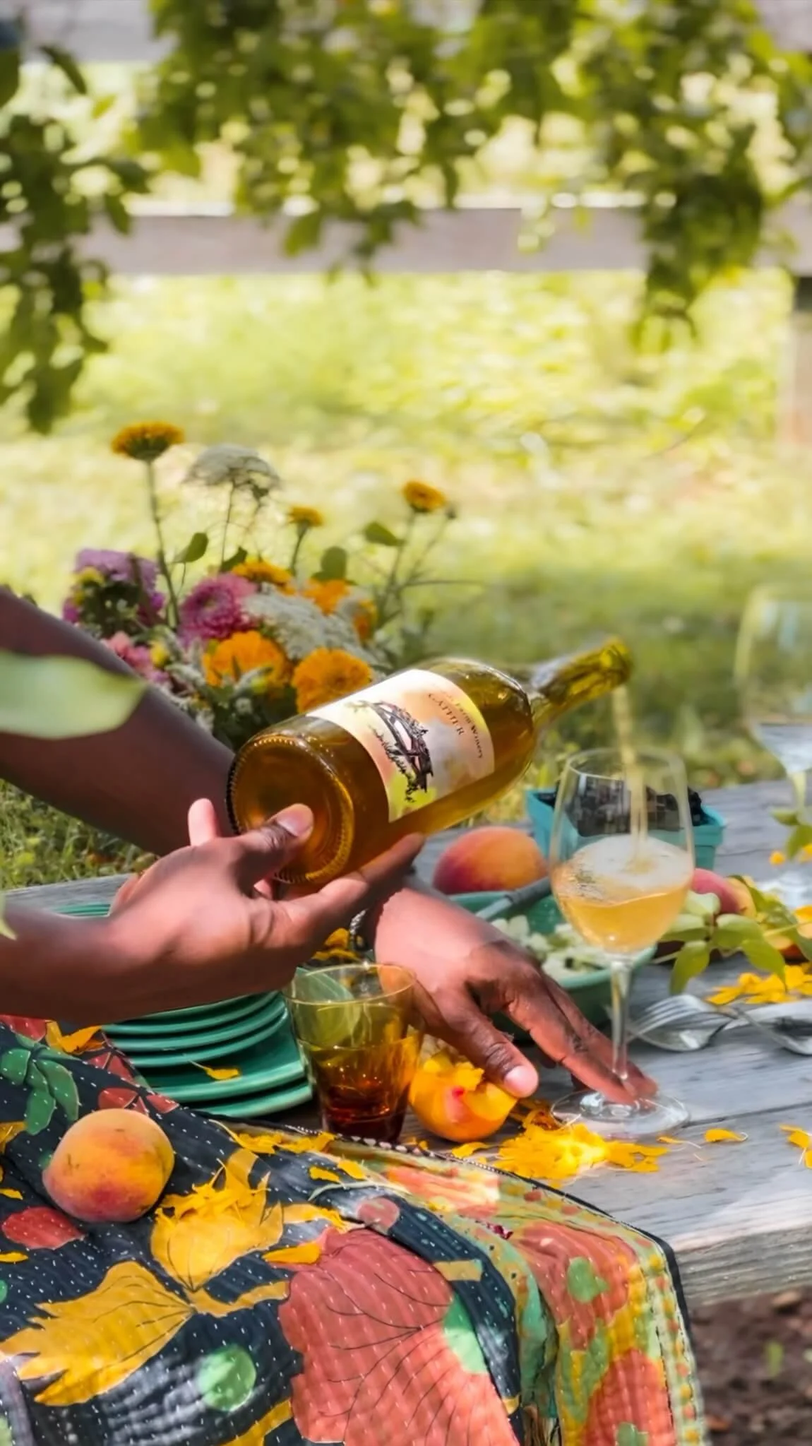 People enjoying a picnic outdoors with flowers, fruit, wine, and colorful tablecloth.