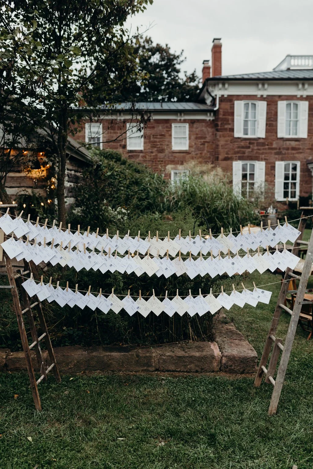 White paper notes attached to clotheslines with clothespins, set up in a garden area in front of a brick house with white window shutters and lush green plants.