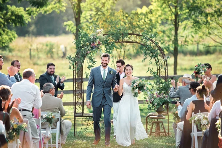 A newlywed couple walking down the outdoor wedding aisle, holding hands, surrounded by guests clapping, with a floral arch and green trees in the background.