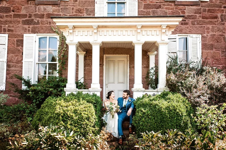 A wedding couple sitting on steps in front of a historic red brick house with white shutters and columns, surrounded by lush green bushes and flowering plants.