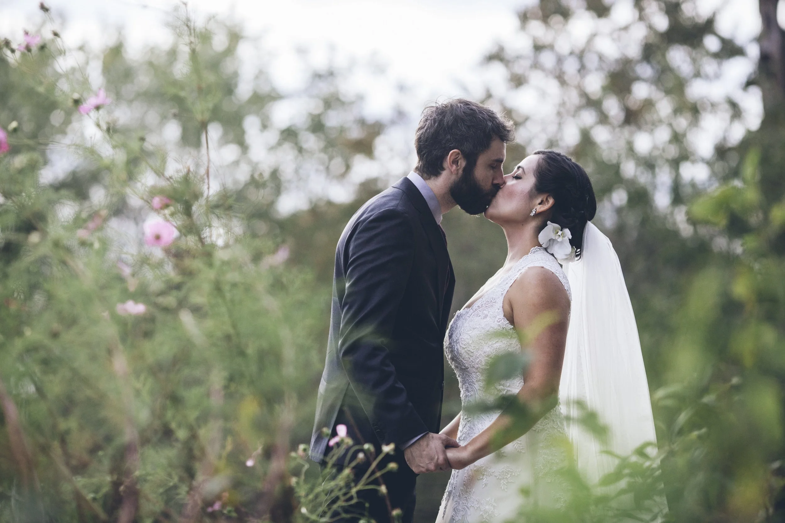 A bride and groom kissing outdoors during their wedding, surrounded by greenery and pink flowers.