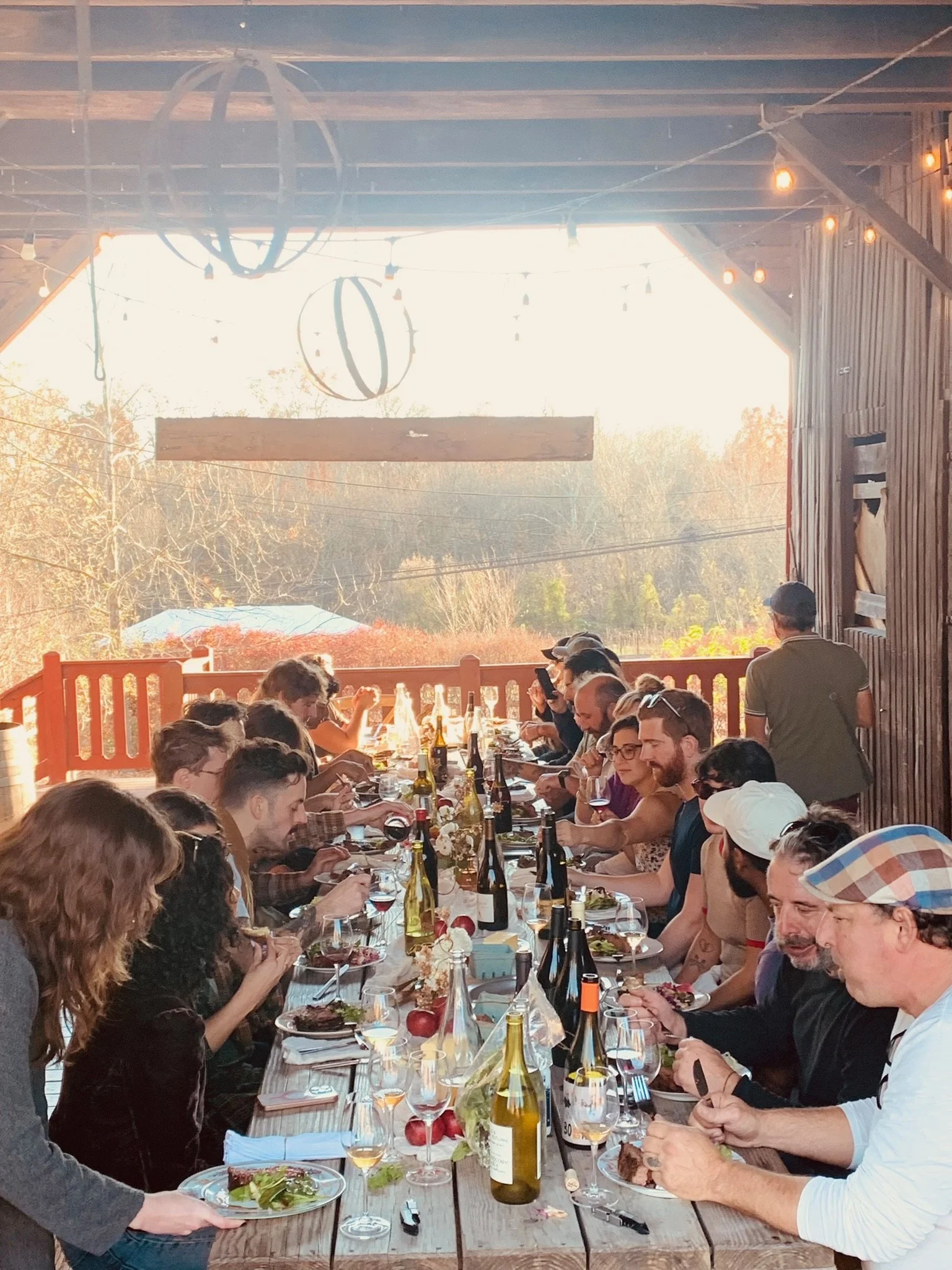 People sitting at a long outdoor table enjoying a meal, with bottles of wine and food dishes, under a wooden roof with string lights, during daytime.