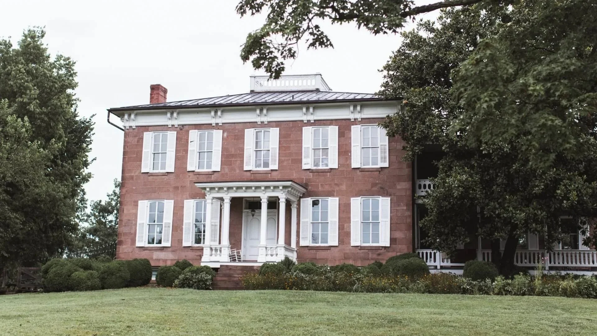 Traditional red brick house with white-shuttered windows, a porch, and a sloped metal roof surrounded by trees and a well-manicured lawn.