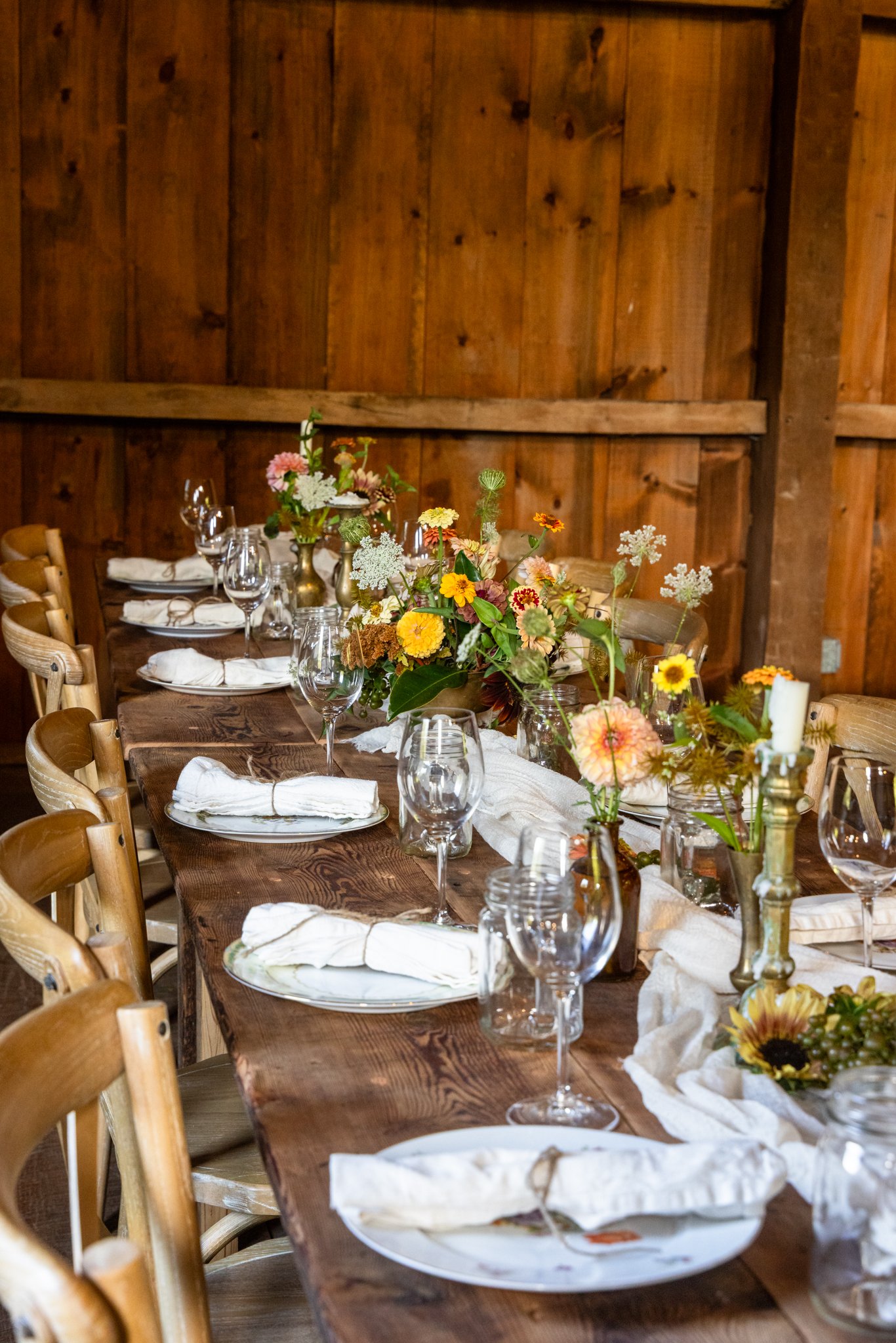 A rustic wooden dining table decorated with a floral centerpiece, surrounded by wooden chairs, with folded white napkins and glassware set for a meal, against a wooden paneled wall.