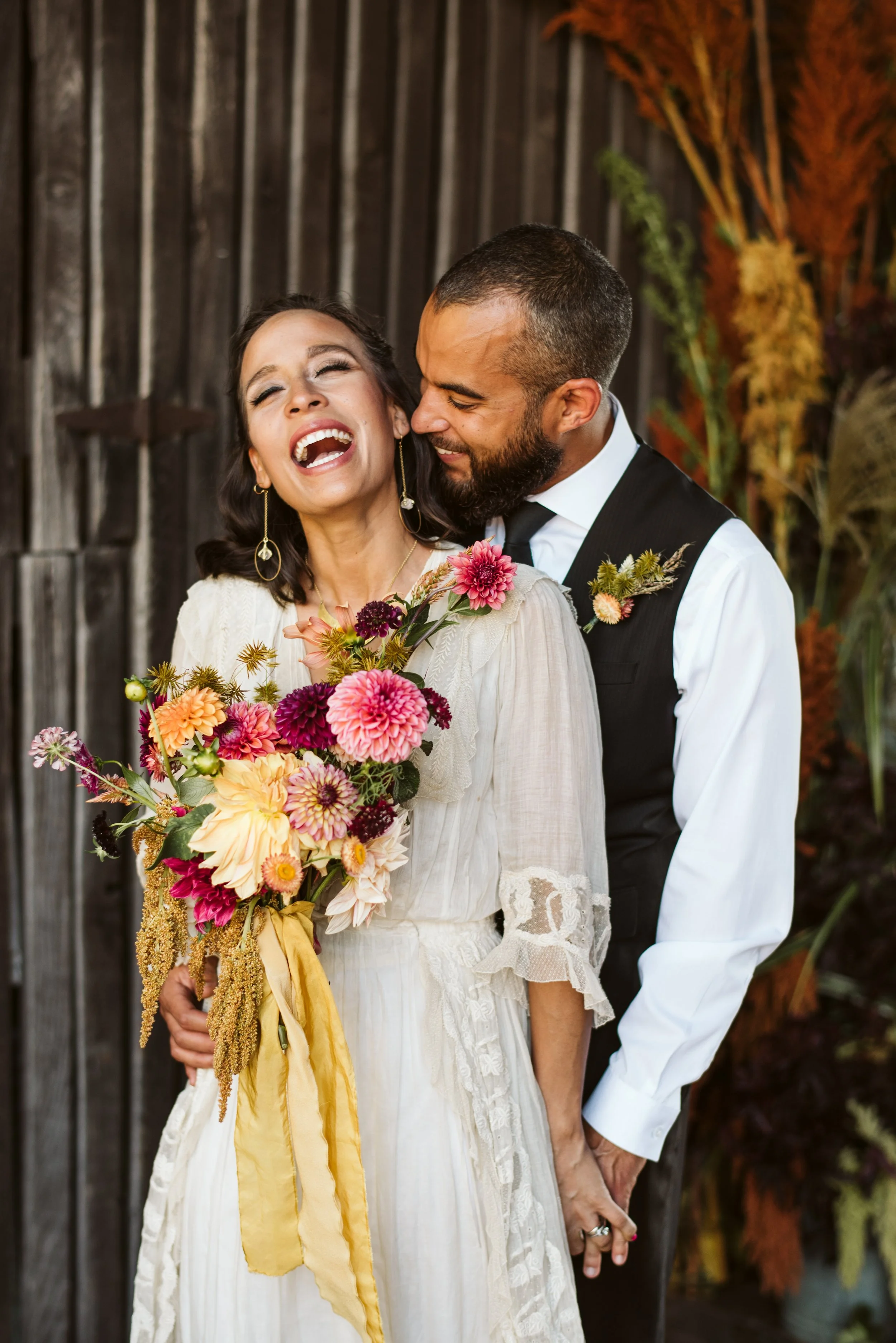 A joyful couple at their wedding, with the woman holding a colorful bouquet of flowers and the man dressed in a vest and white shirt, standing outdoors against a wooden fence with autumnal foliage.