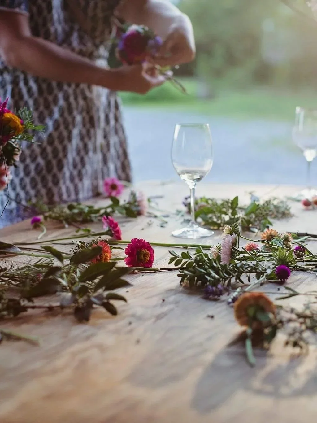 A woman in a patterned dress arranging flowers on a wooden table outdoors, with empty wine glasses and scattered floral decorations.