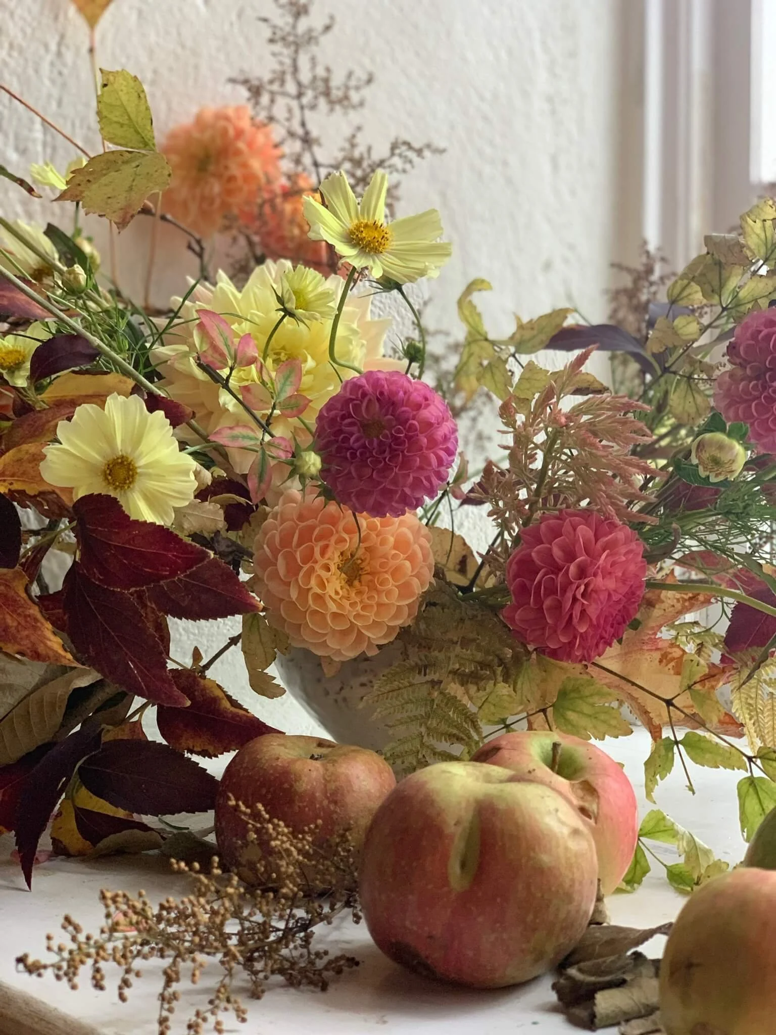 A still life arrangement of apples, various flowers including dahlias and cosmos, dried leaves, and branches on a white surface near a window.