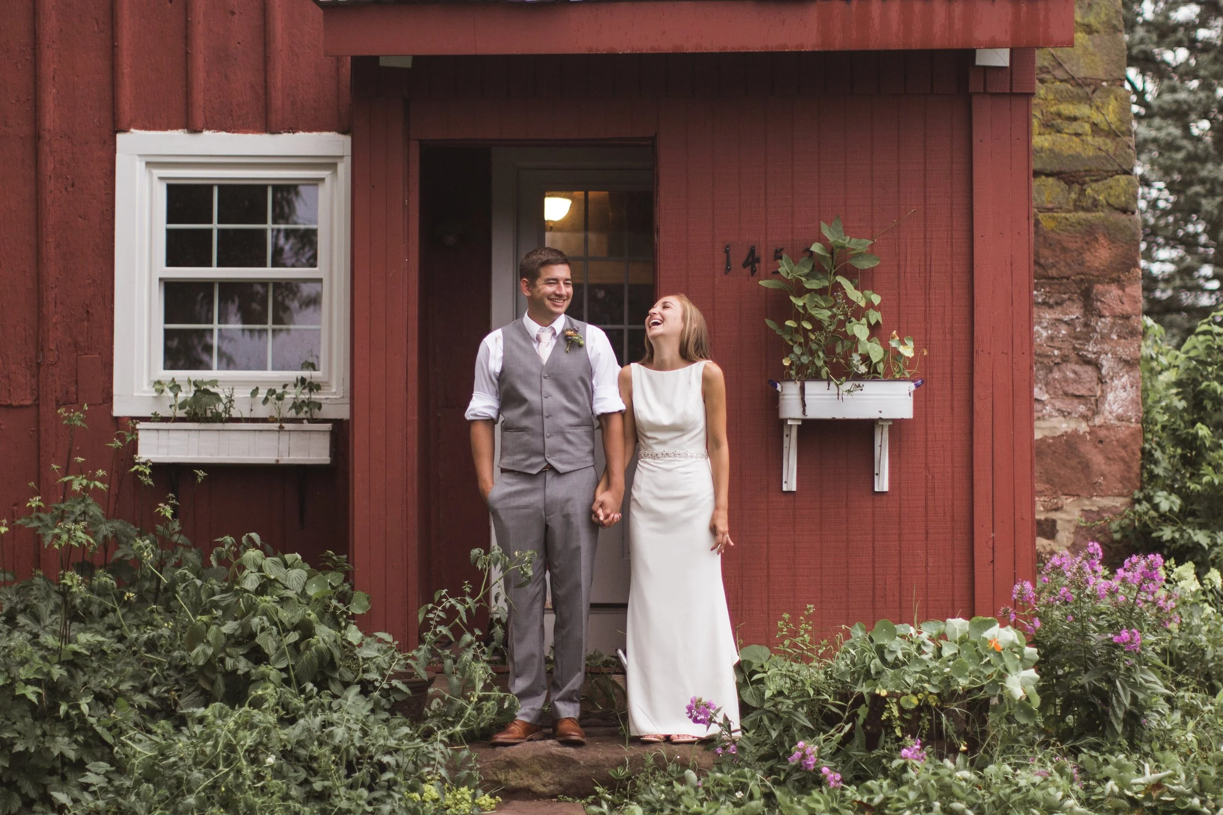 A couple dressed in wedding attire standing outside a red wooden house, holding hands and laughing together.
