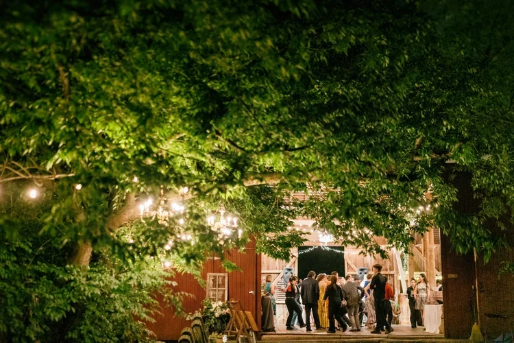 People dancing and socializing in a rustic barn decorated with string lights, surrounded by trees.