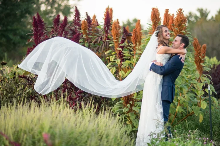 A newlywed couple in wedding attire standing outdoors in a garden with colorful plants; the bride's long veil is flowing in the wind, and the groom is lifting the bride as they smile at each other.
