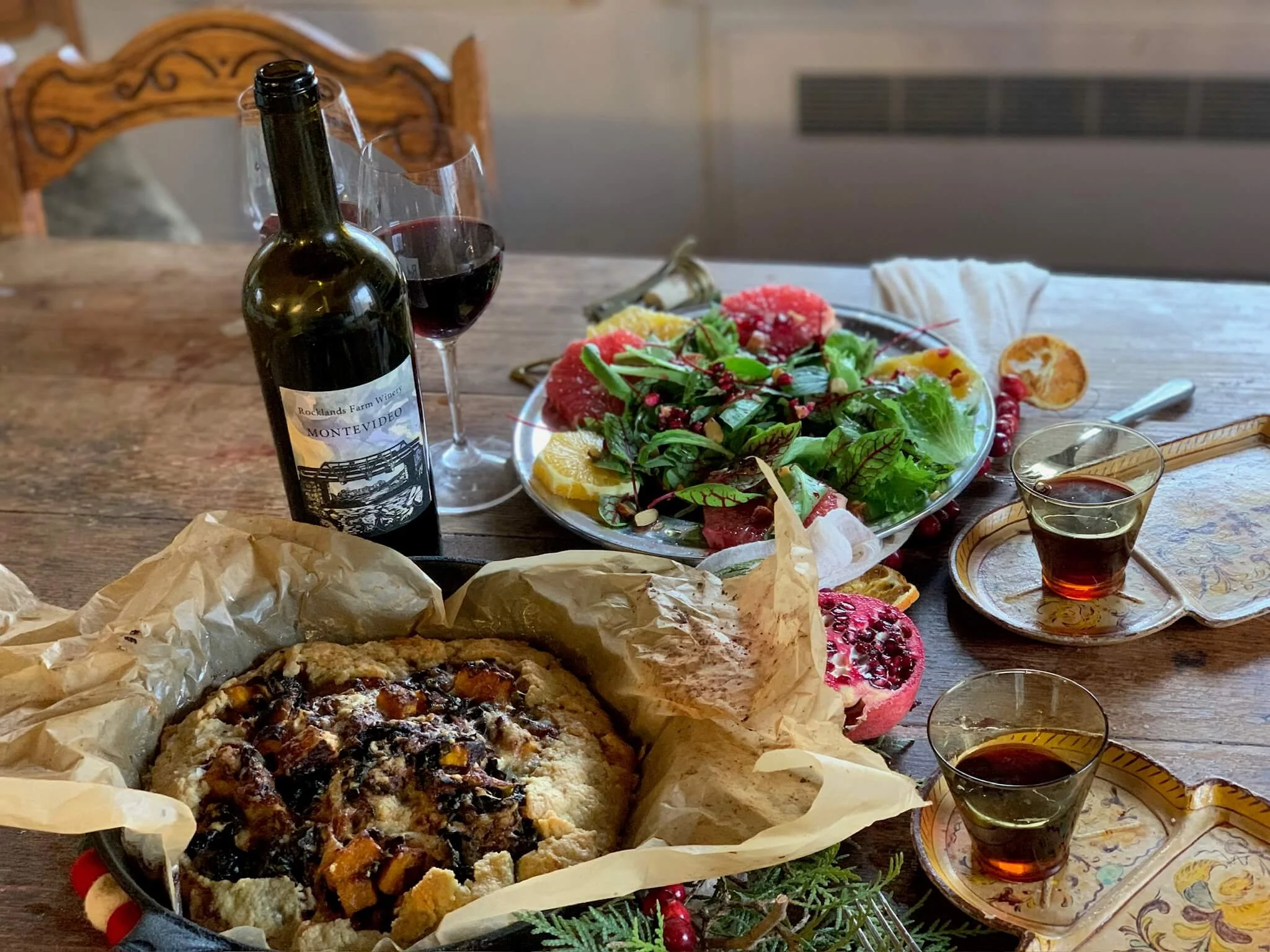 A table set with a bottle of red wine, a glass of red wine, a fresh salad with greens and citrus slices, a partially eaten pie or dessert on parchment paper, and small glasses of amaretto or similar liquor. The background includes wooden chairs and a
