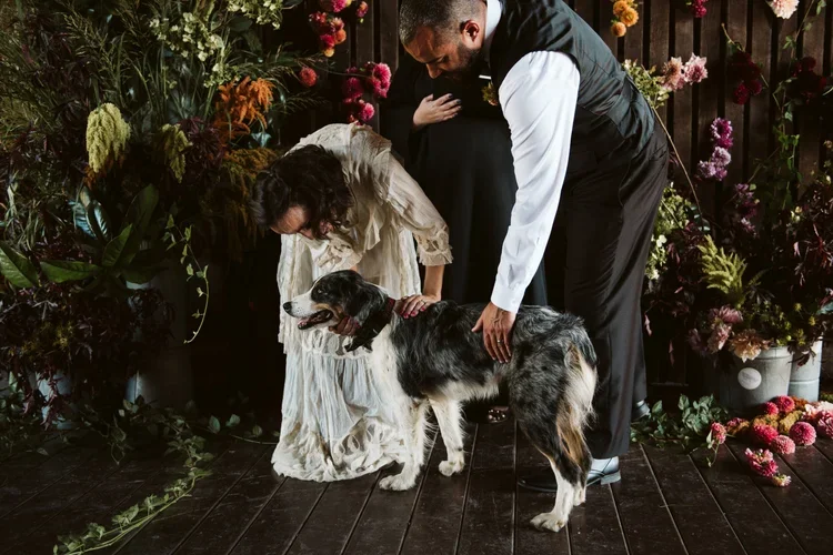 A couple with a dog at a wedding or special event, standing on a dark wooden floor surrounded by colorful flowers and greenery.