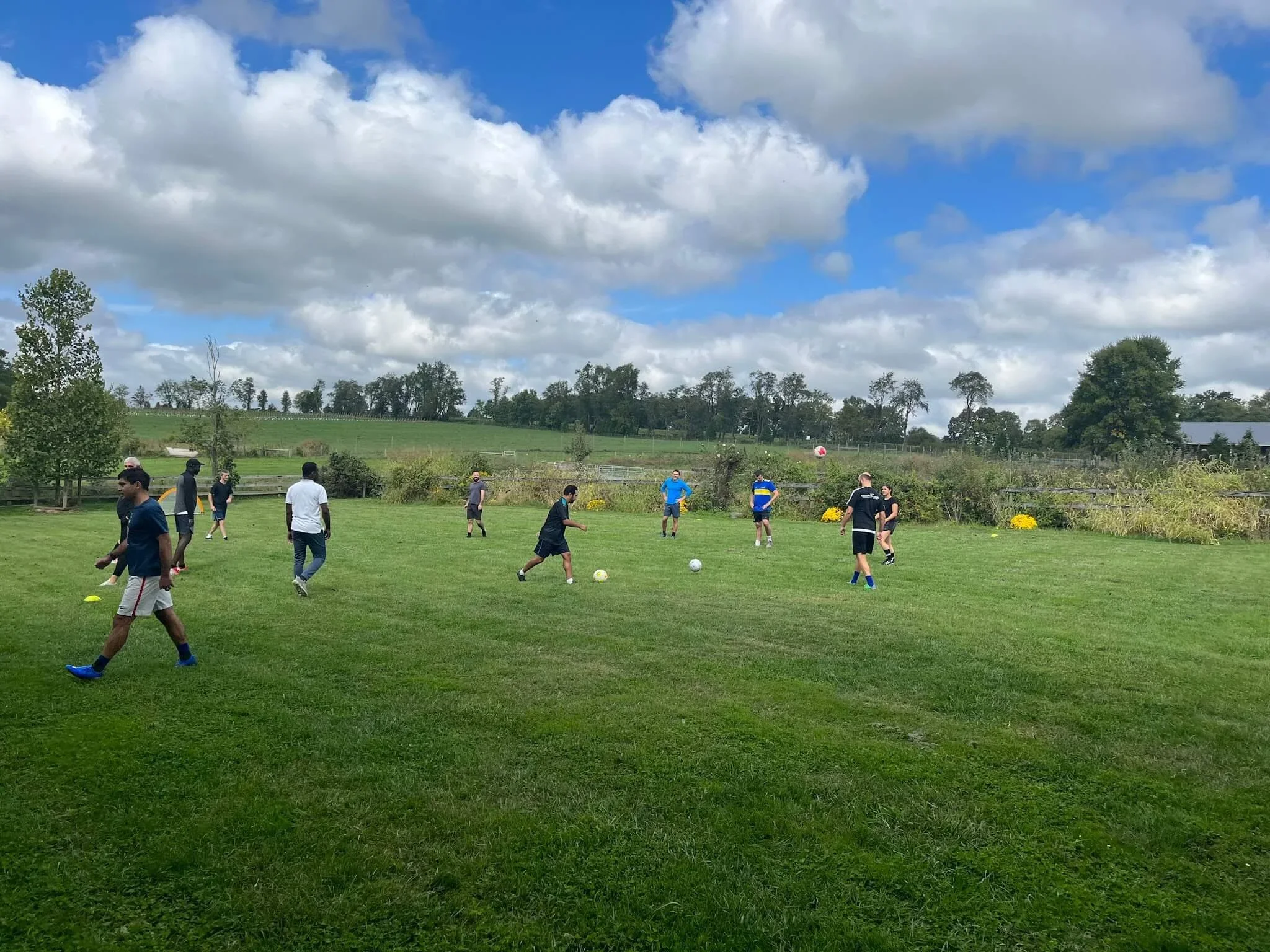 Group of children and teenagers playing soccer on a grassy field outdoors under a partly cloudy sky, with trees and rolling hills in the background.