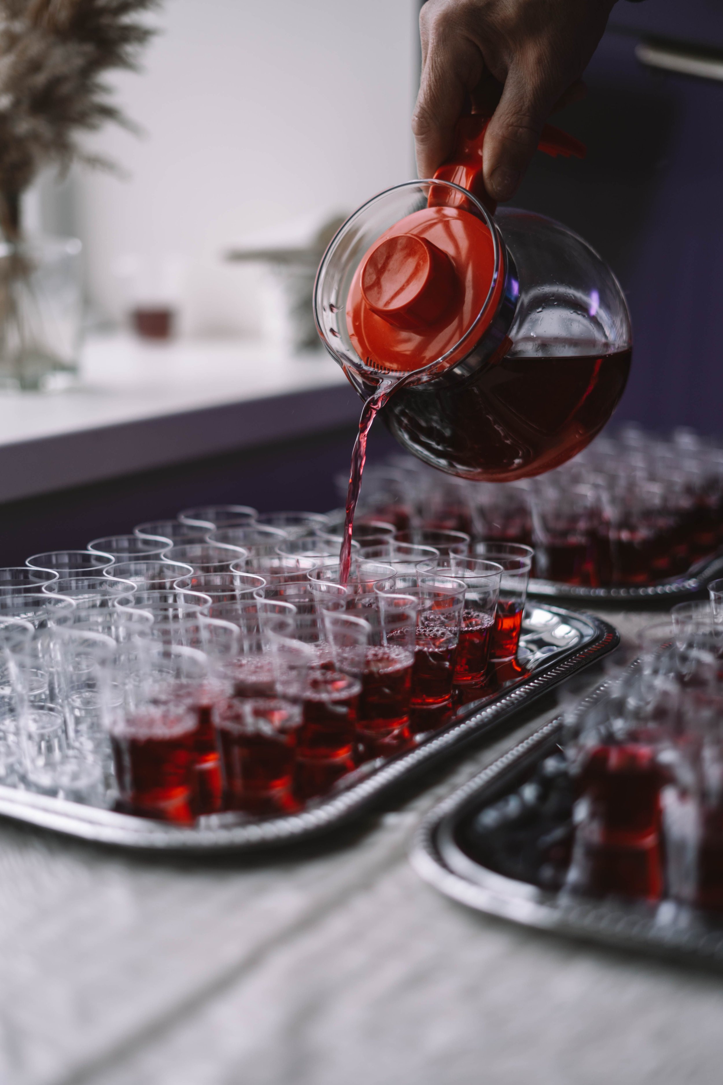A person pouring red liquid from a glass pitcher into small clear shot glasses arranged on a tray.