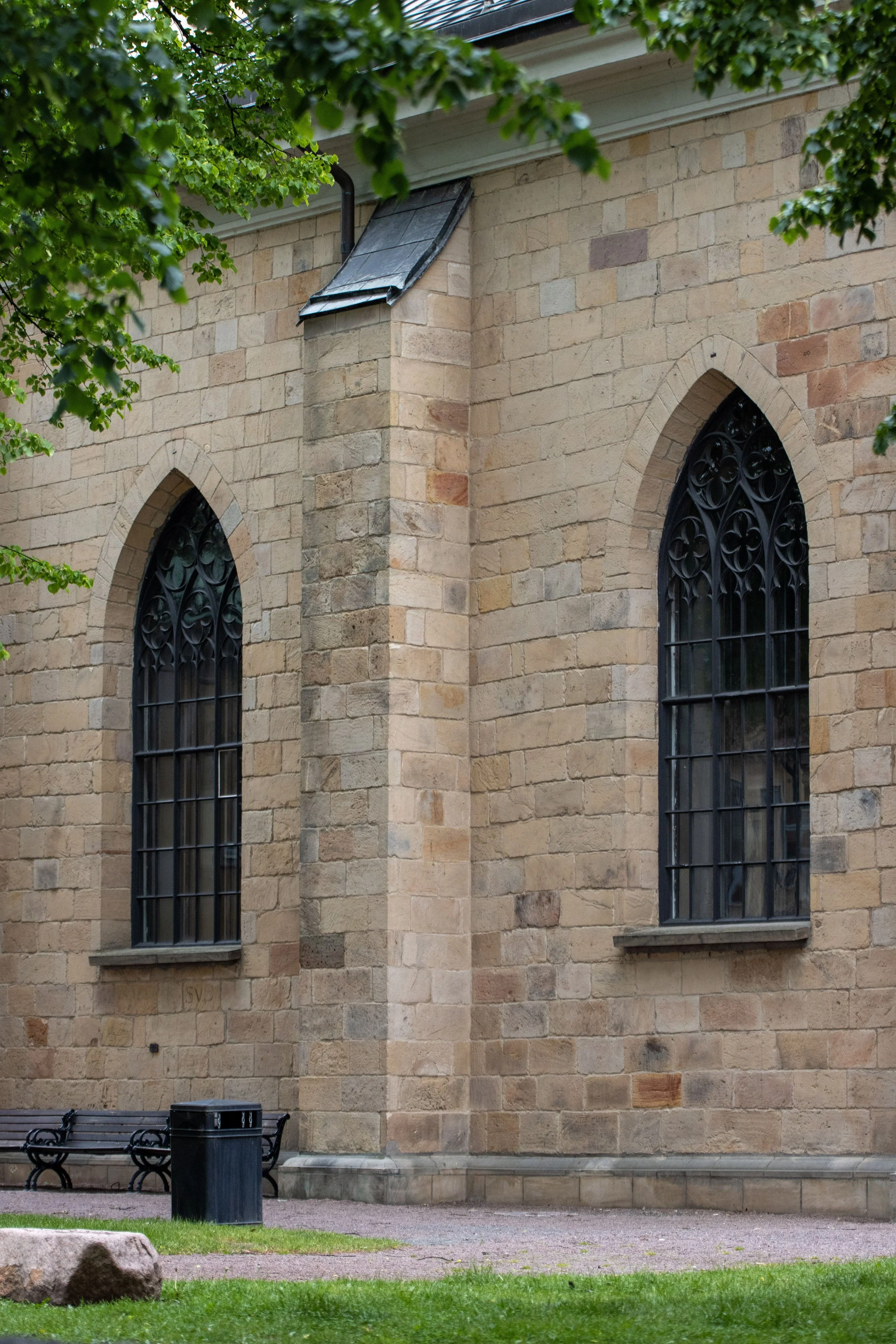 A section of the exterior of a stone church with large, pointed arch windows with decorative black iron grilles. Green leaves partially frame the top of the image. A bench and trash can are on the ground near the building.