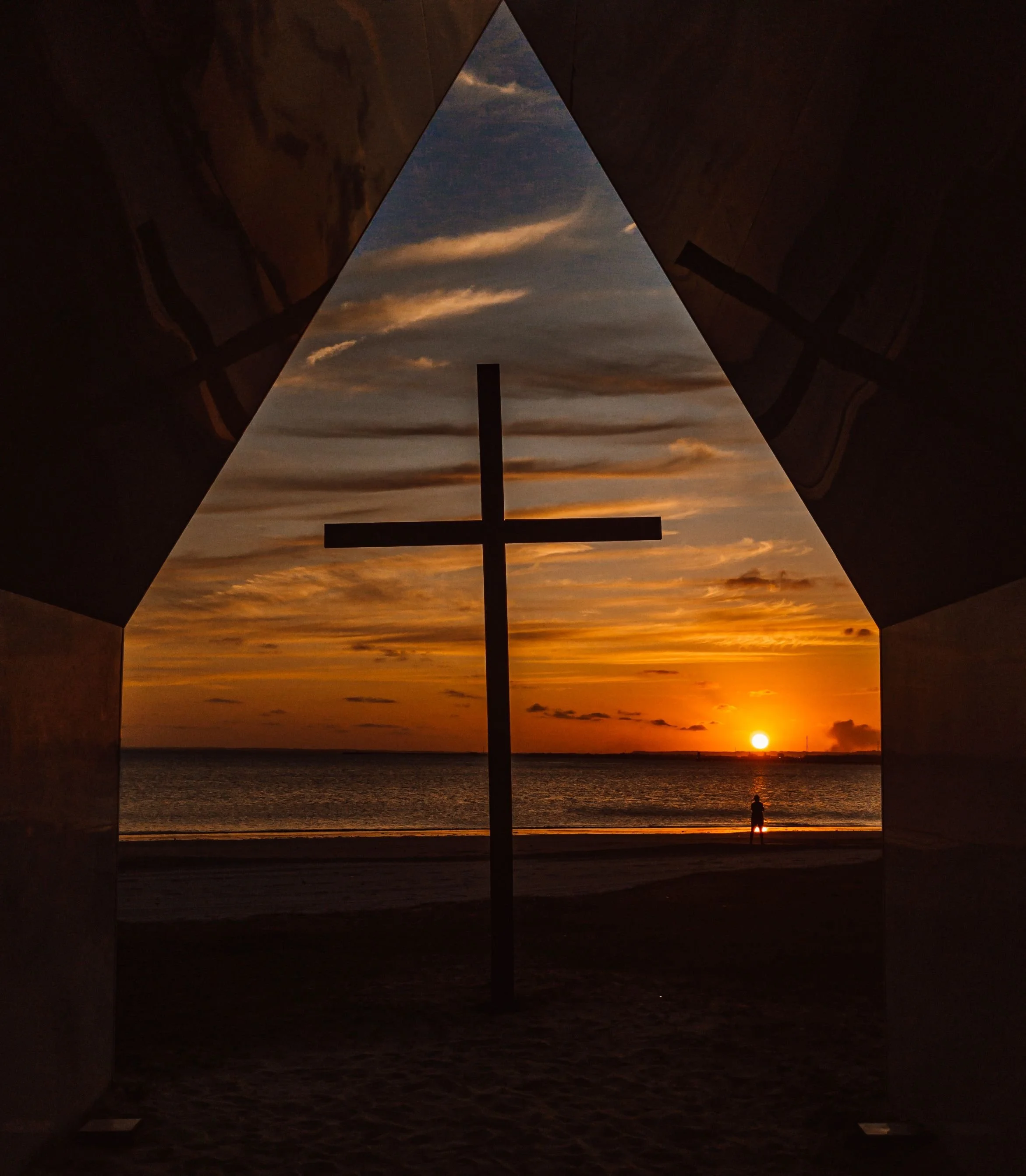 View of a sunset over the ocean seen through an open symmetrical architectural structure with dark walls, a cross in the center, and a person standing on the beach.