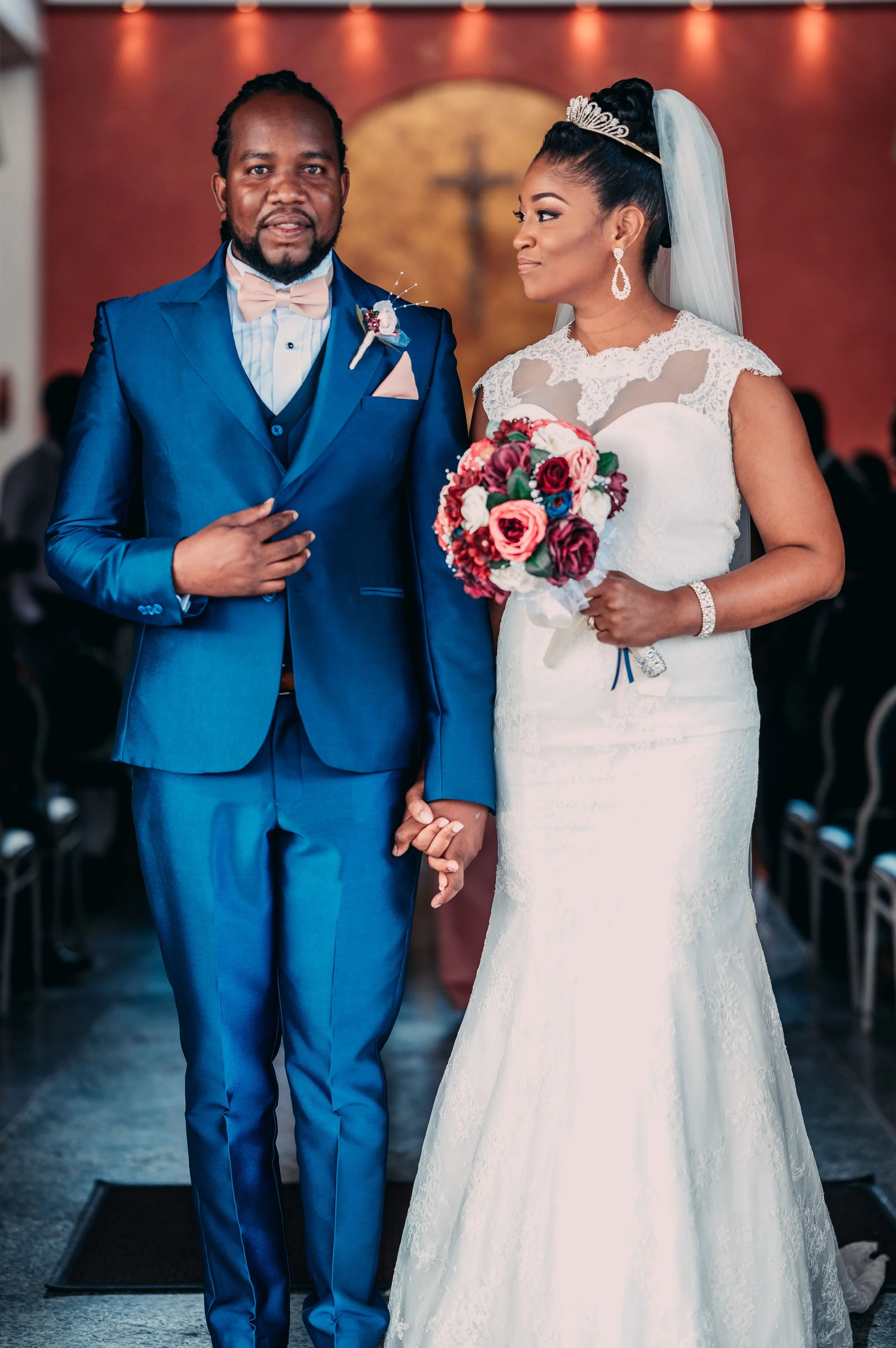 A couple dressed in wedding attire standing hand-in-hand inside a church. The man wears a blue suit, light pink bow tie, and a boutonniere. The woman wears a white lace wedding dress, veil, tiara, and holds a bouquet of red, pink, and white flowers. They are looking at each other.