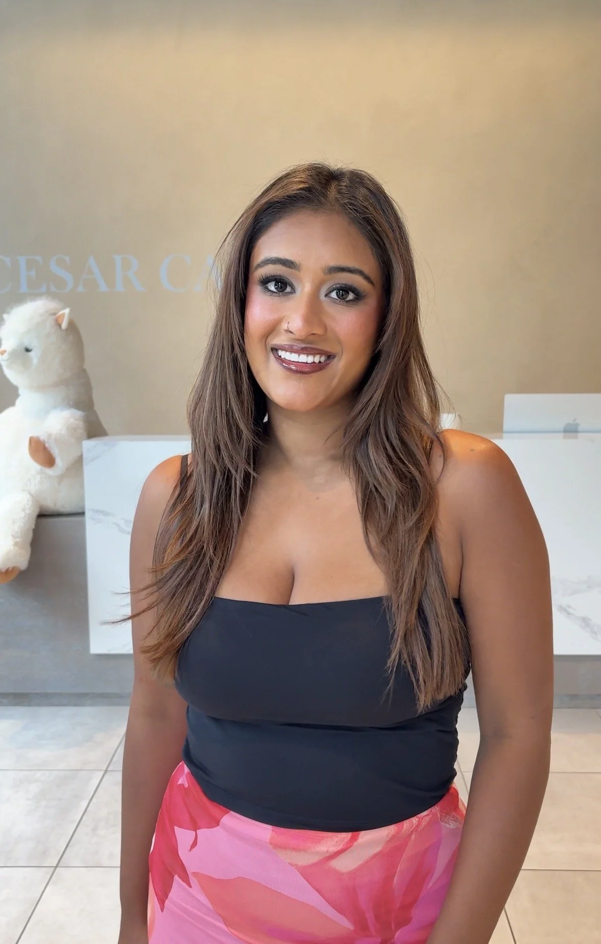 A smiling woman with long brown hair, wearing a black top and a pink patterned skirt, standing indoors in front of a beige wall with a white bear plush toy in the background.