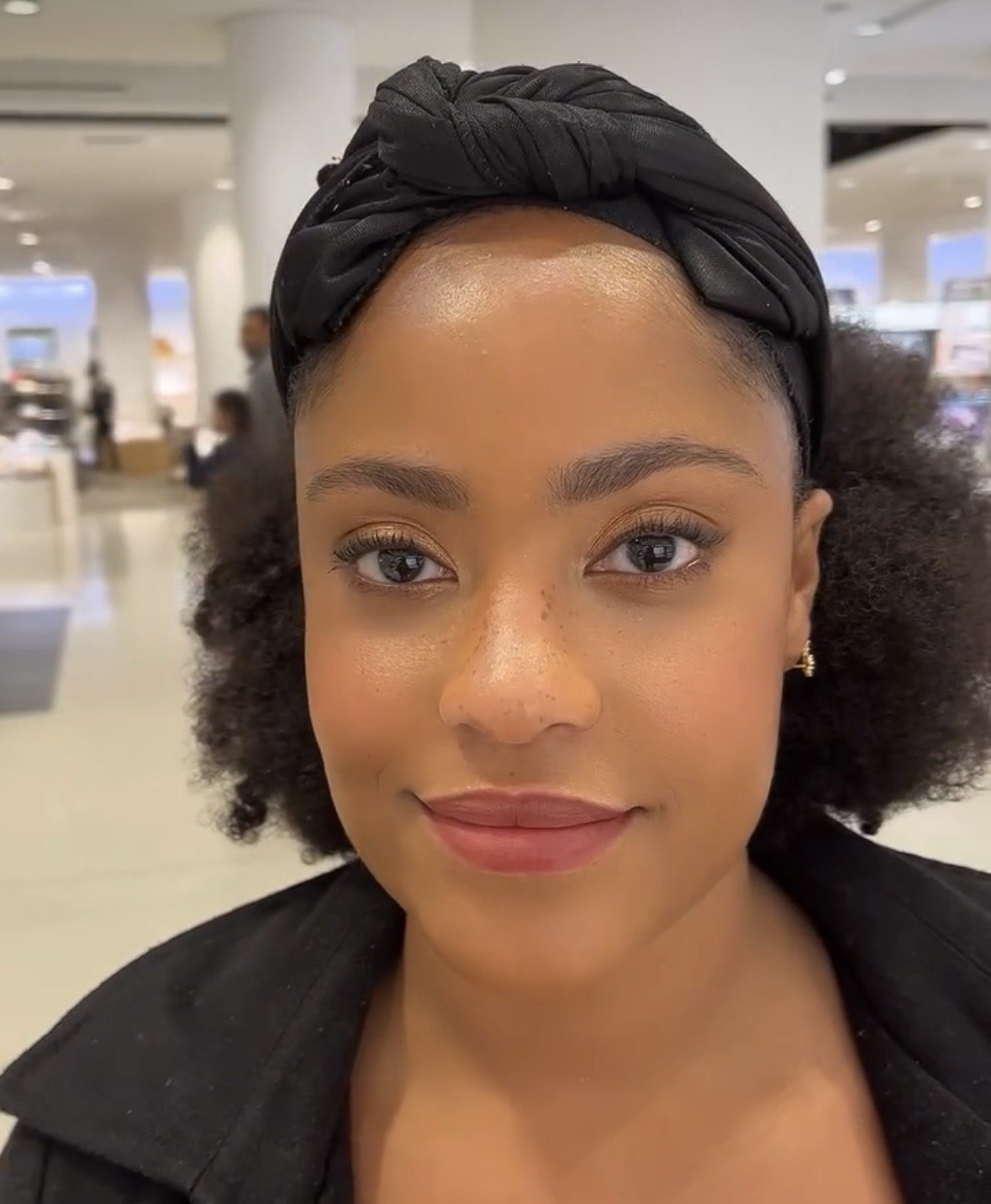 A close-up of a woman with natural curly hair, wearing a black headscarf, and smiling inside a store or mall.