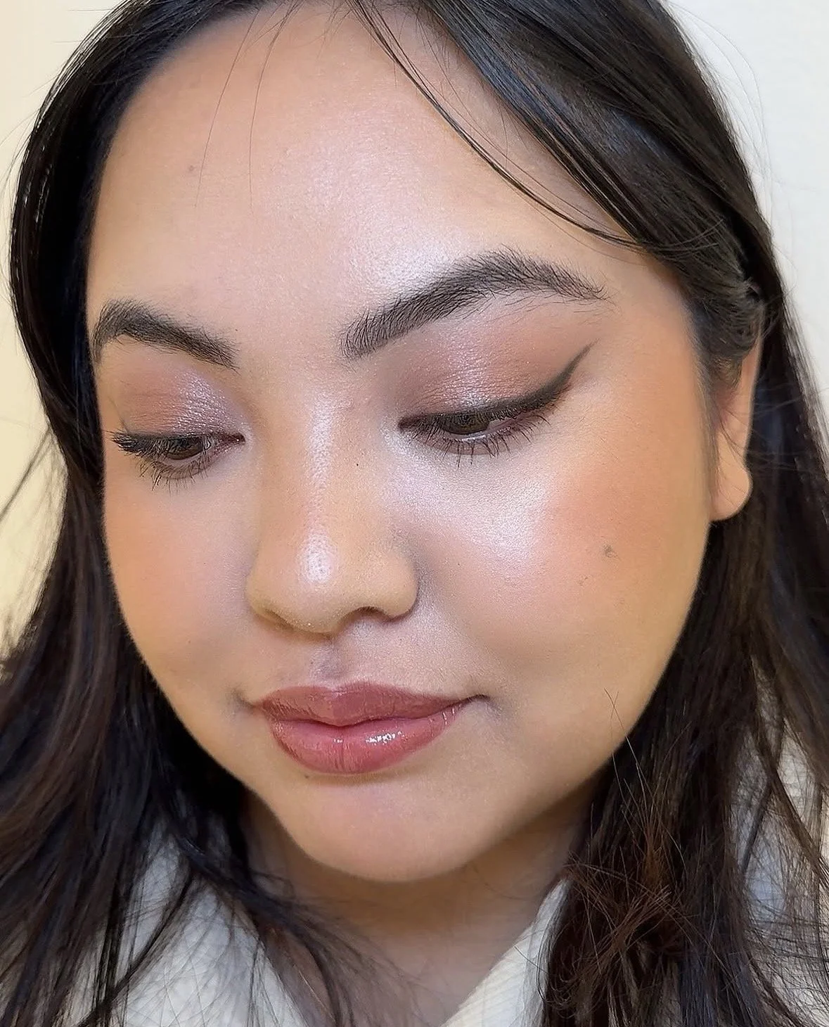 A young woman with dark hair and makeup, looking down with closed eyes, showcasing her eye makeup and pink lip gloss.