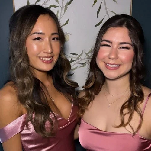 Two women smiling and posing for a photo, both wearing pink satin dresses and having wavy hair, standing in front of a decorative wall with green leaf patterns.