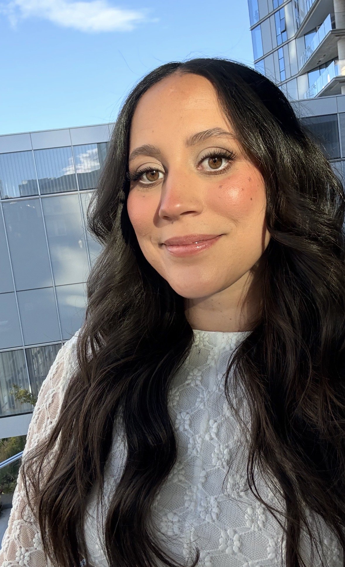 A woman with long dark hair and brown eyes taking a selfie outdoors with a glass building and a blue sky in the background.