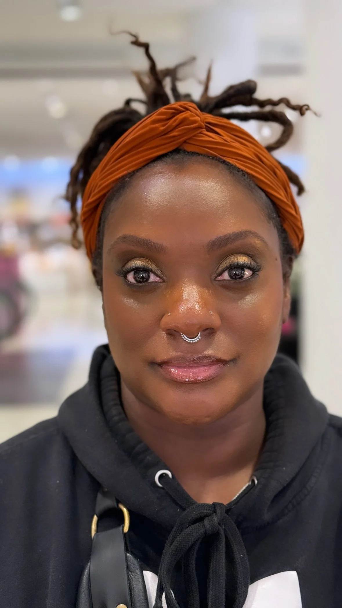 A woman with medium brown skin and dreadlocks wearing an orange head wrap, a septum piercing, and a black hoodie, standing indoors with a blurred background.